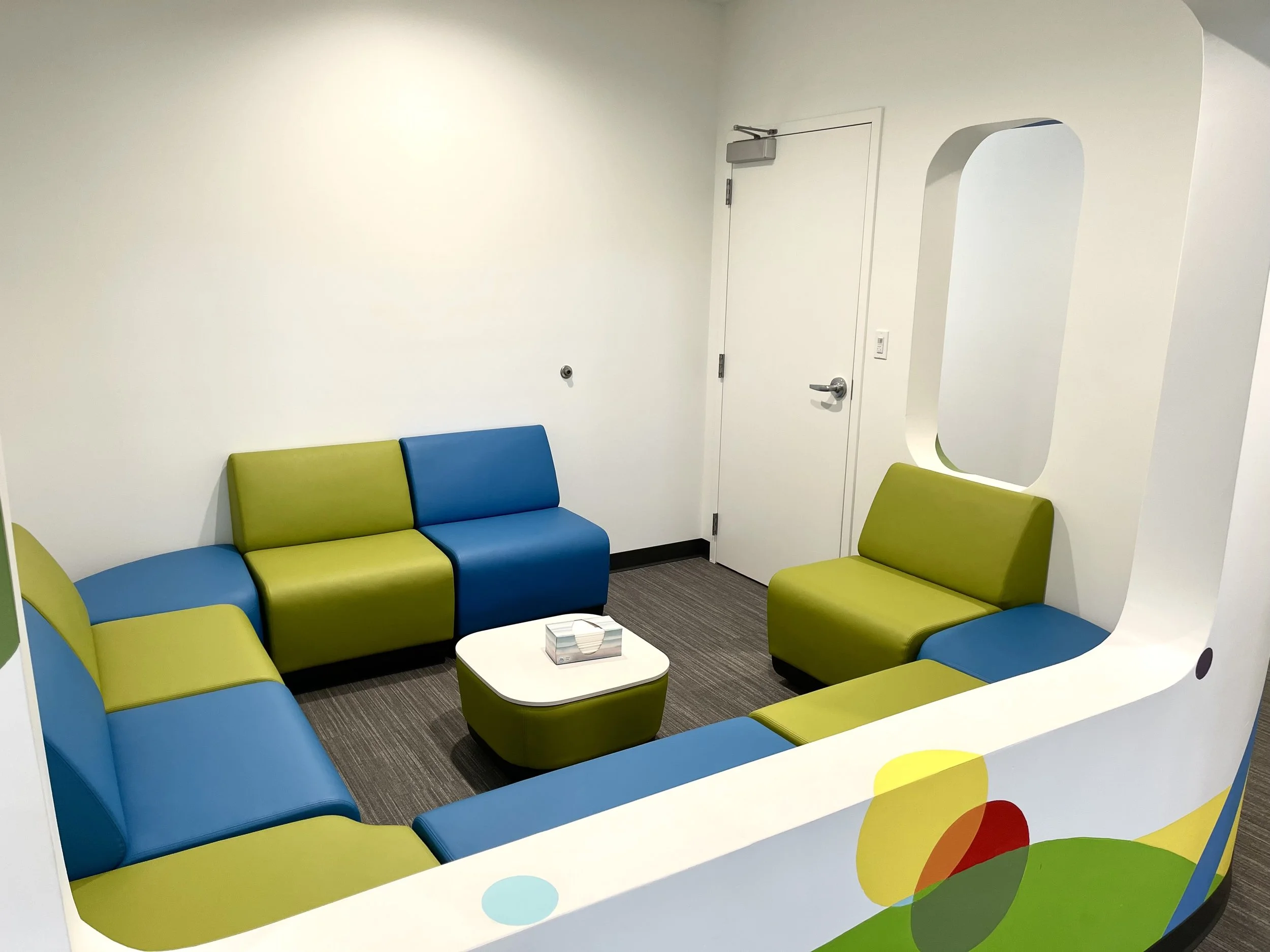 A colorful waiting area with blue, green, and yellow cushioned chairs arranged in a semi-circle around a small white table. The space has white walls, a door, and an arched opening with a mirror. There is a box of tissues on the table.