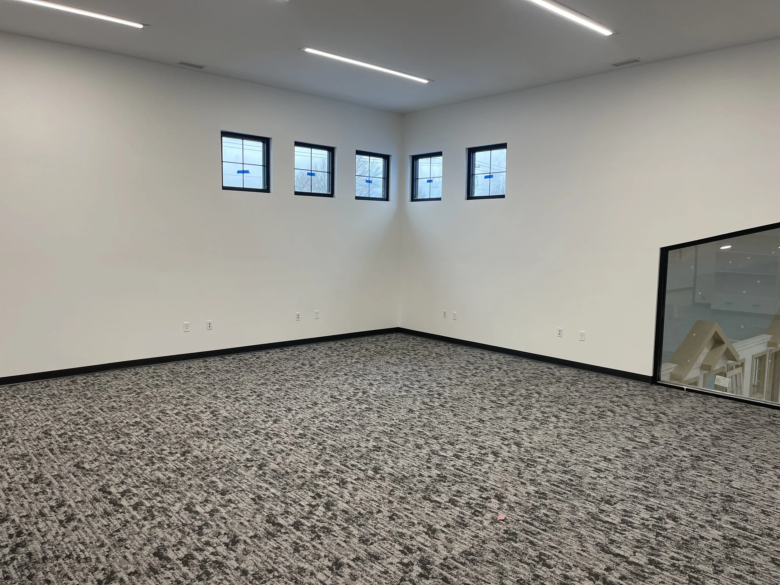 Empty interior room with white walls, multiple windows near the ceiling, gray and black patterned carpet, and a corner with a glass wall panel.