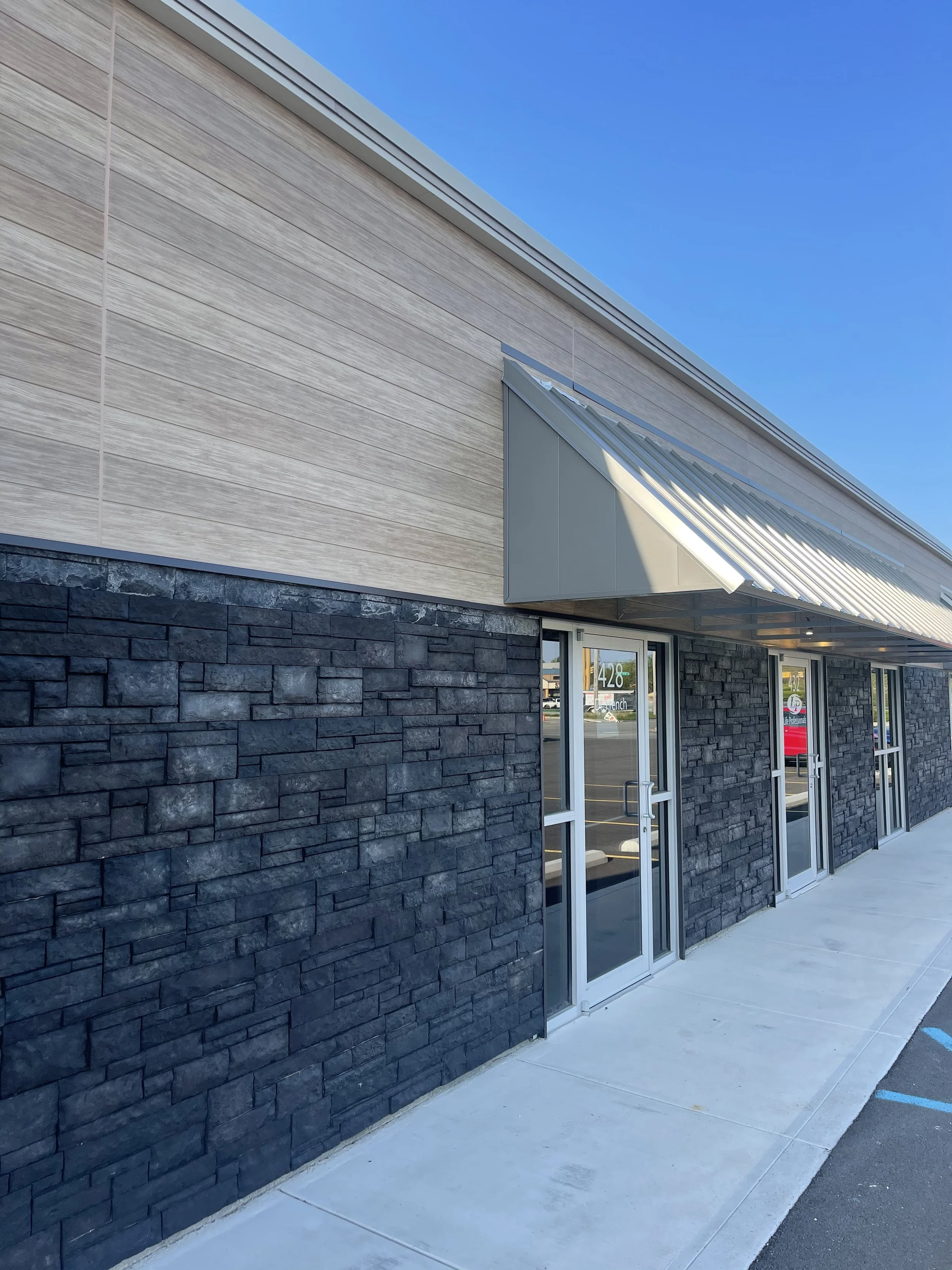 Exterior of a modern building with a black stone facade, wood siding, and large glass doors under a partly cloudy sky.