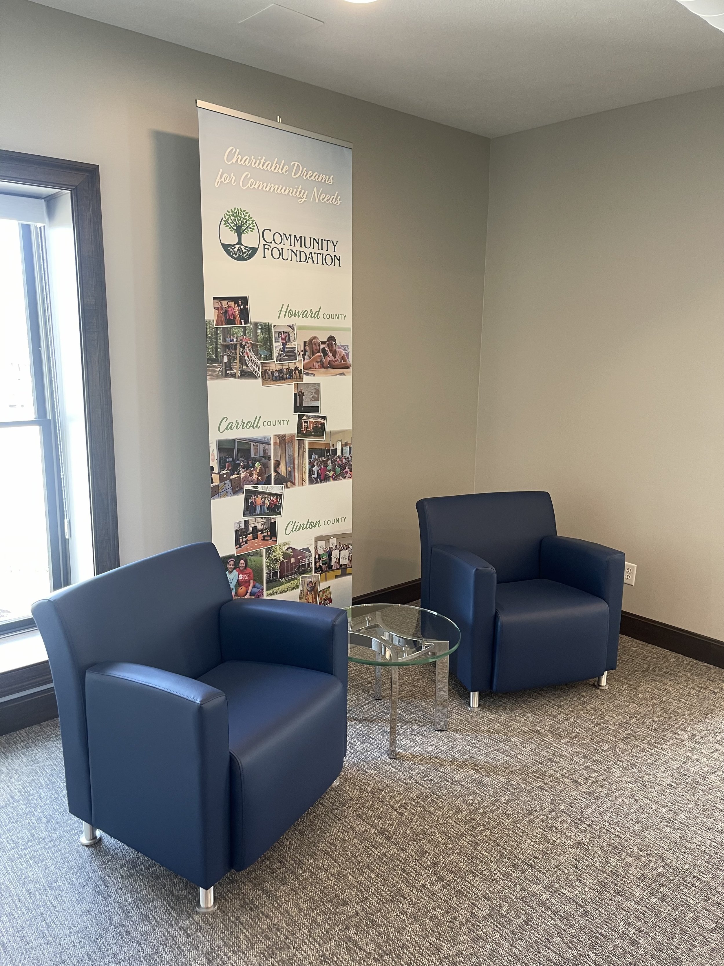 A waiting area with two dark blue armchairs, a round glass table, and a vertical banner displaying information about a community foundation. The room has beige carpet, a gray wall, a window with sunlight, and an electrical outlet.