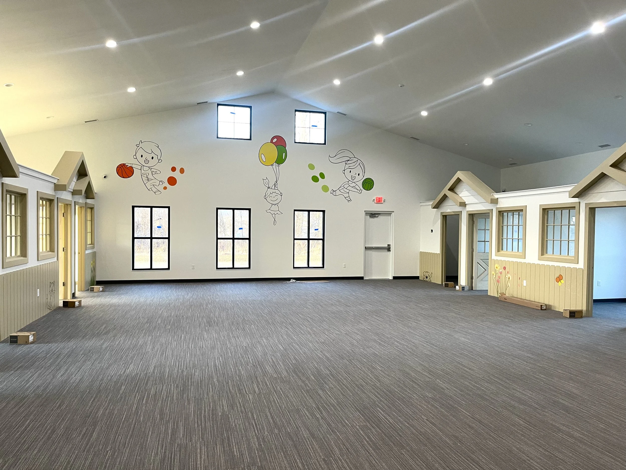 Interior of a brightly lit children’s playroom with mural art of children playing with sports balls and balloons on the white walls, and small house-shaped structures with windows and flower decorations.