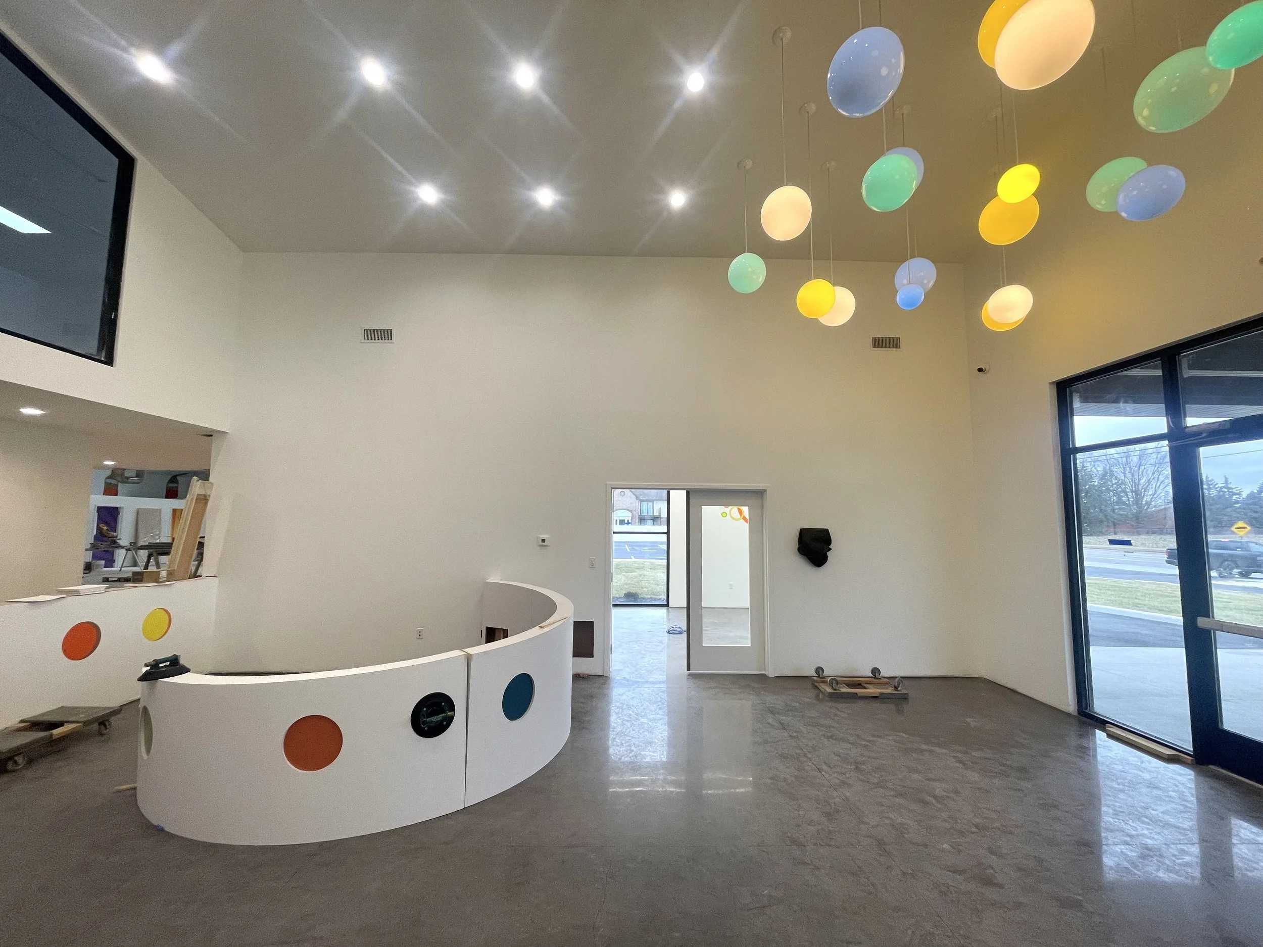 Interior of a building under construction or renovation with white walls, large windows, a curved white reception desk with multicolored dots, and a ceiling with recessed lighting and hanging colorful spherical lights.