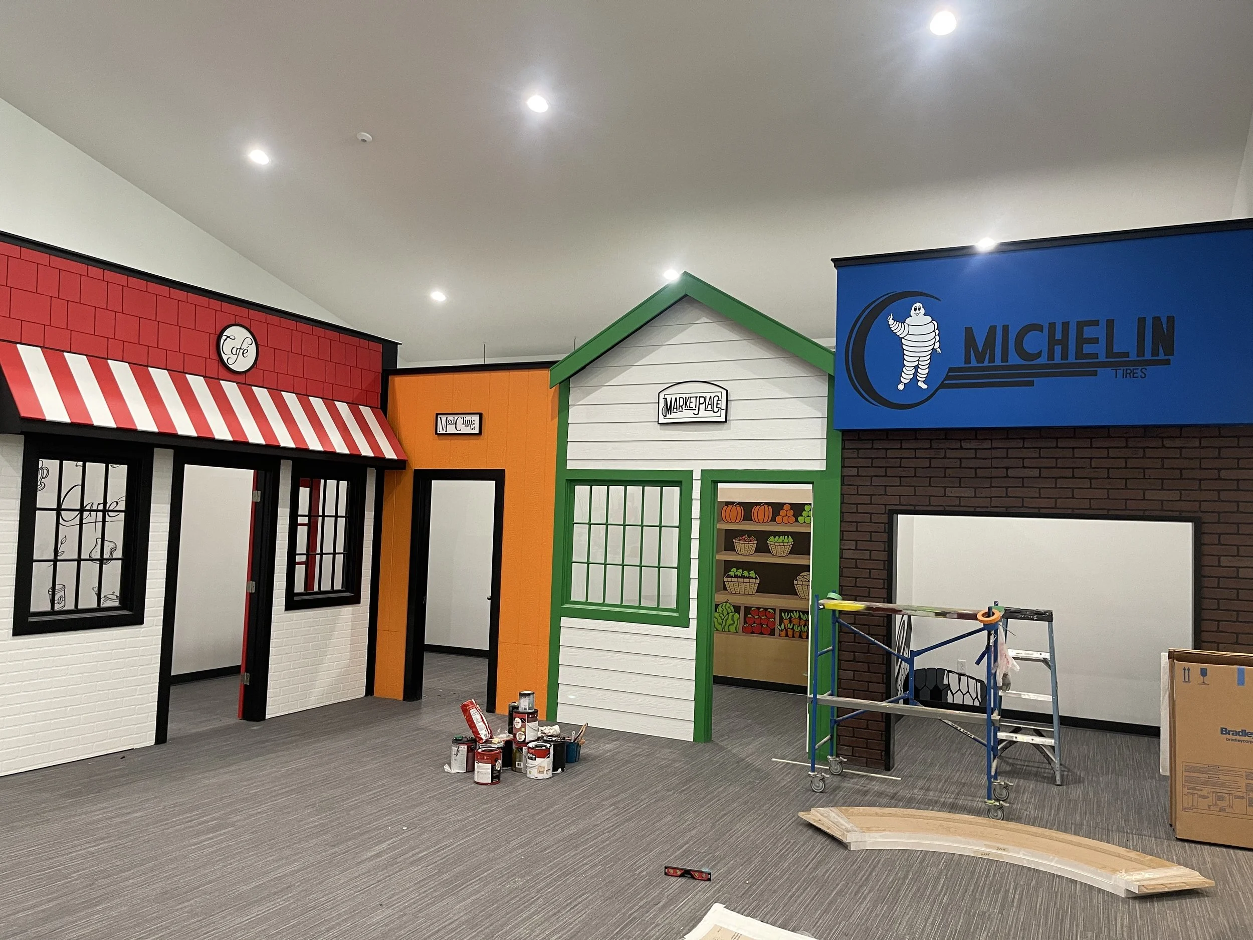 Indoor space under construction or renovation with colorful small buildings facade, paint cans, boxes, scaffolding, and an unfinished floor.