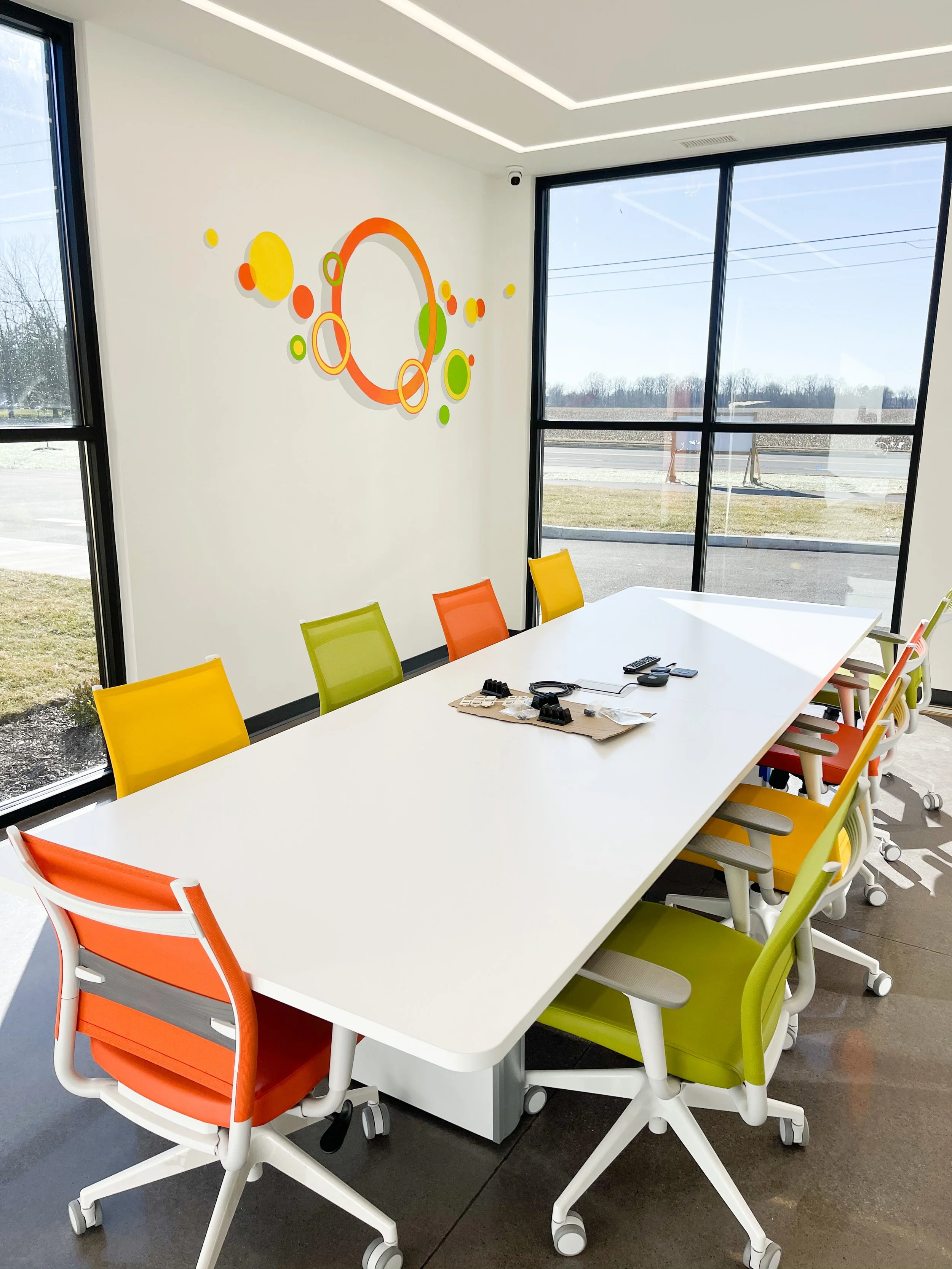 Modern office conference room with colorful chairs around a white table, large windows with a view of the outdoors, and abstract wall art featuring circles in yellow, orange, and green.