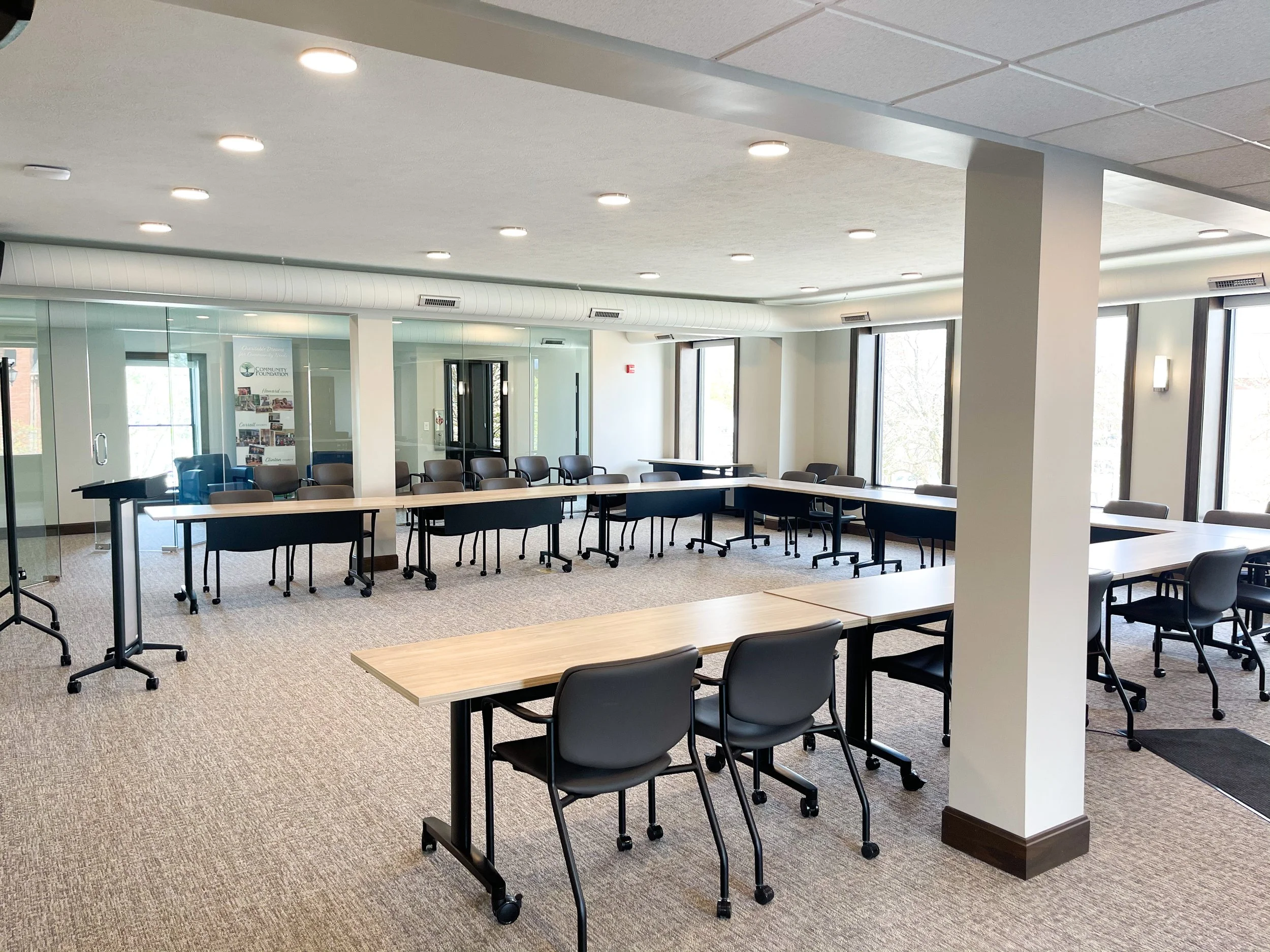 An empty conference room with tables and chairs, large windows, ceiling lights, and light-colored walls.
