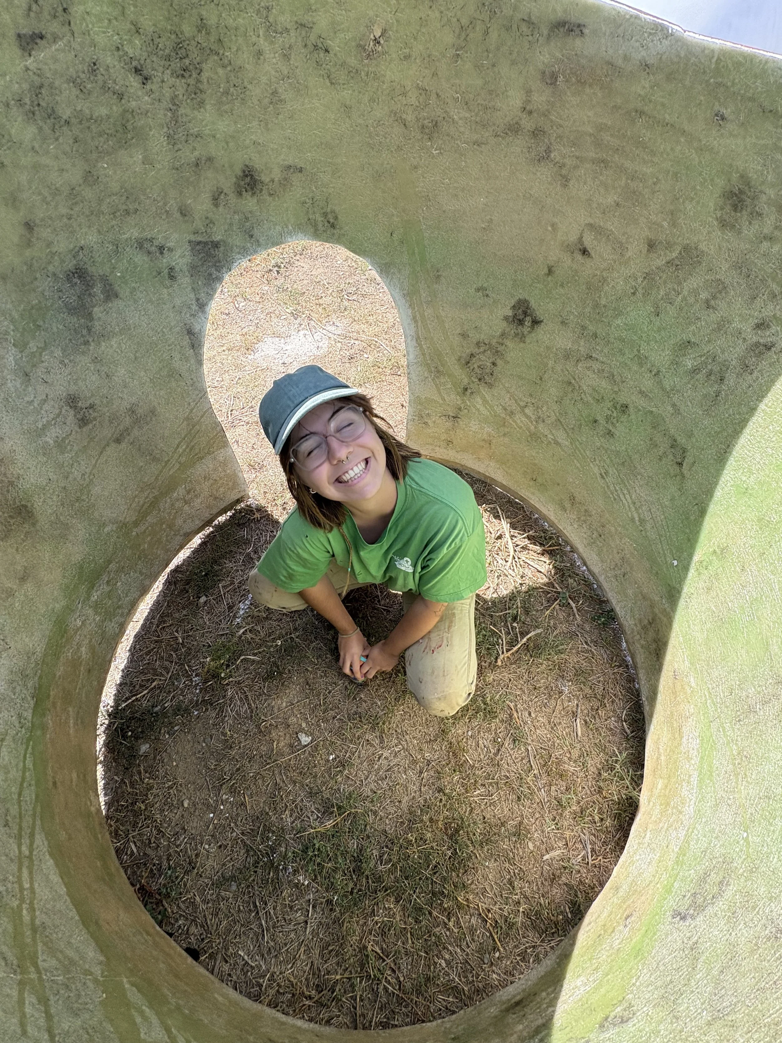A smiling person with glasses and a hat kneeling inside a hollow, large, greenish, cylindrical sculpture outdoors.