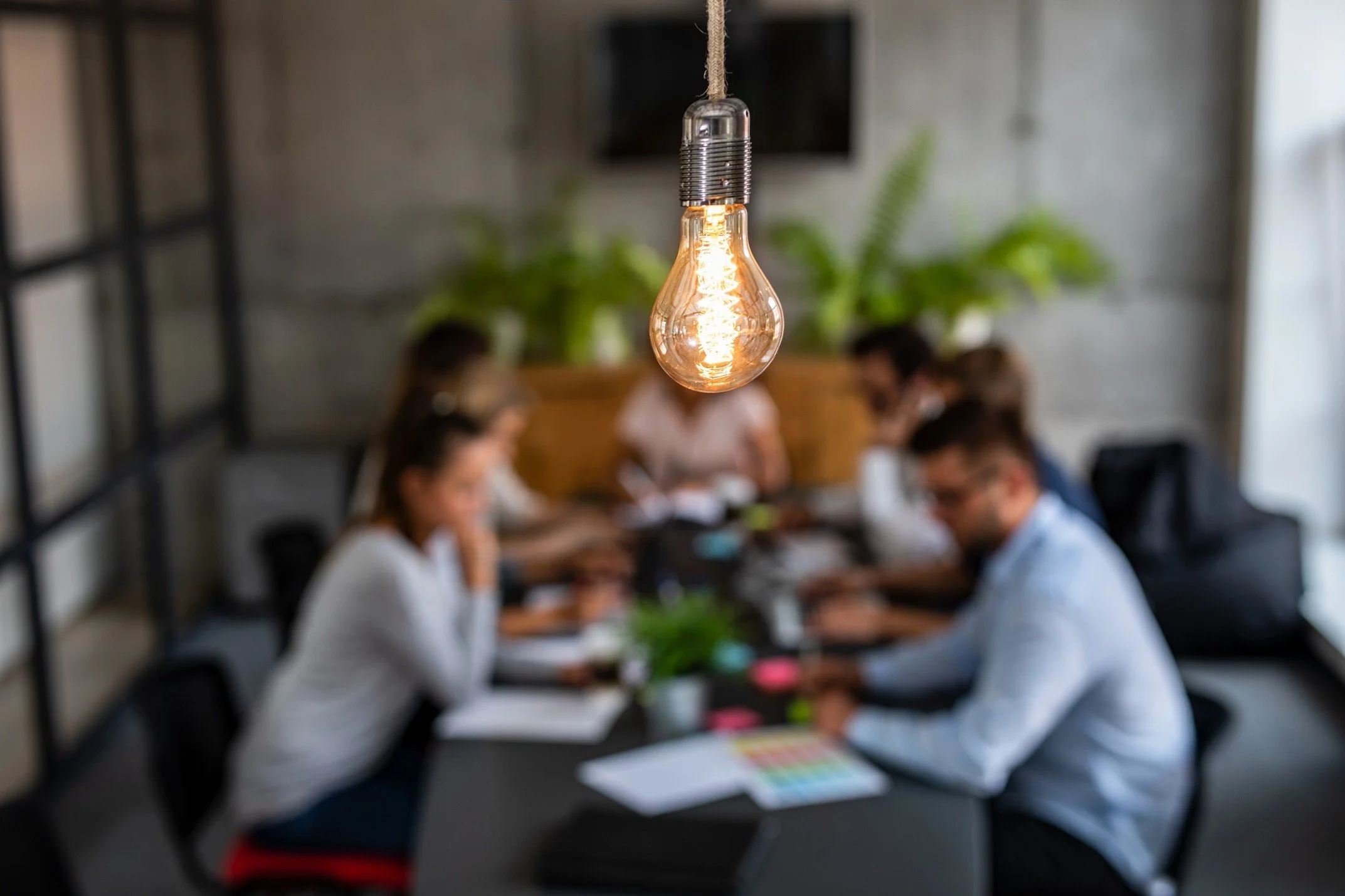 A hanging Edison-style light bulb with a glowing filament in focus, with blurred people sitting at a table in a modern indoor setting in the background.