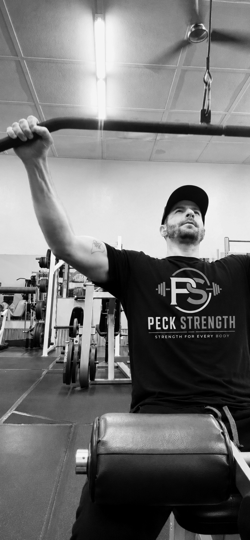 A man at the gym pulldown weights while seated, wearing a black t-shirt with 'Peck Strength' logo, in a Mariposa, CA gym with weight racks and equipment in the background.