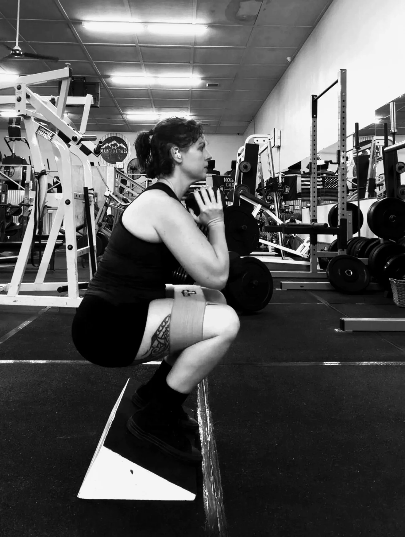 A woman performing a squat exercise with a weight on her shoulders inside a Mariposa, CA gym, with equipment and weights visible in the background.