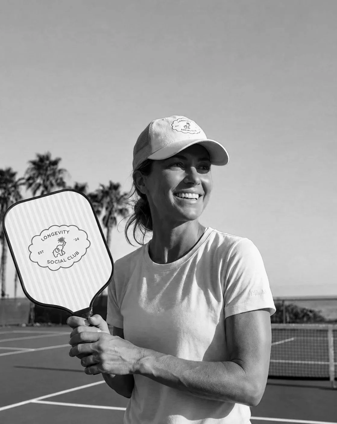 Smiling woman holding a paddle with 'Longevity Social Club' logo on a tennis court in black and white