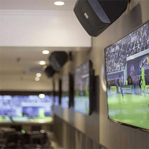 Multiple flat-screen TVs mounted on a wall in a room, displaying a televised sports event, with speakers overhead and lighting on the ceiling.
