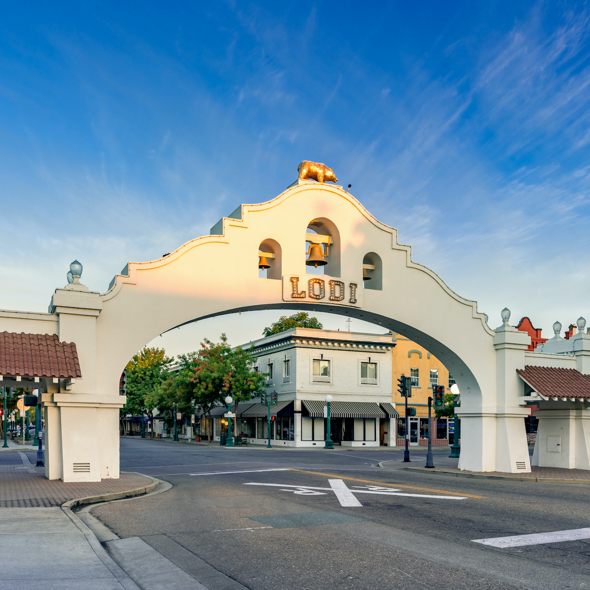Street scene viewed through an archway with the word 'Lodi' and a bell, against a partly cloudy sky.