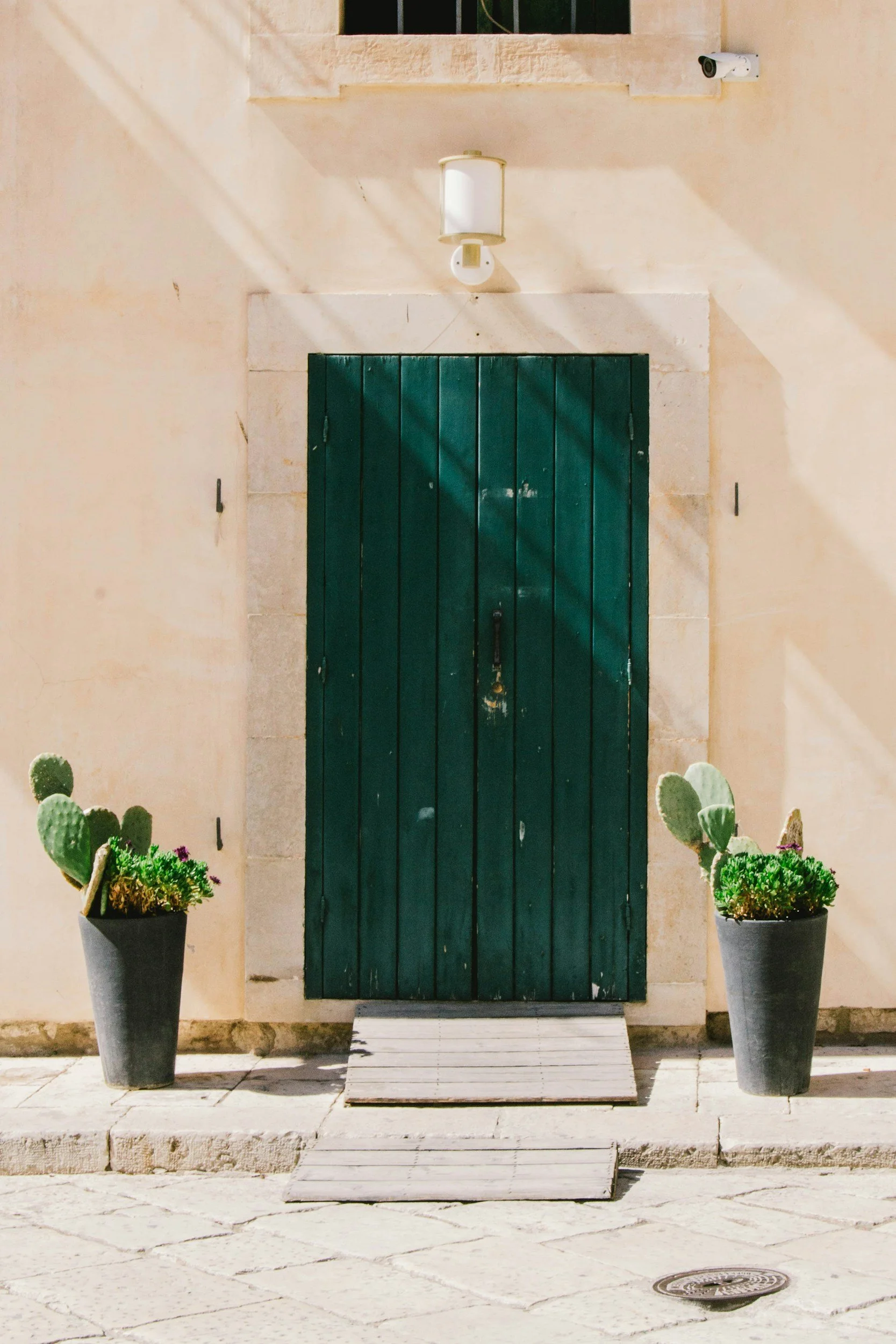 A closed dark green wooden door with a small white doormat in front, flanked by two potted cactus plants, against a light-colored stucco wall, with a security camera on the upper right and a small barred window above.
