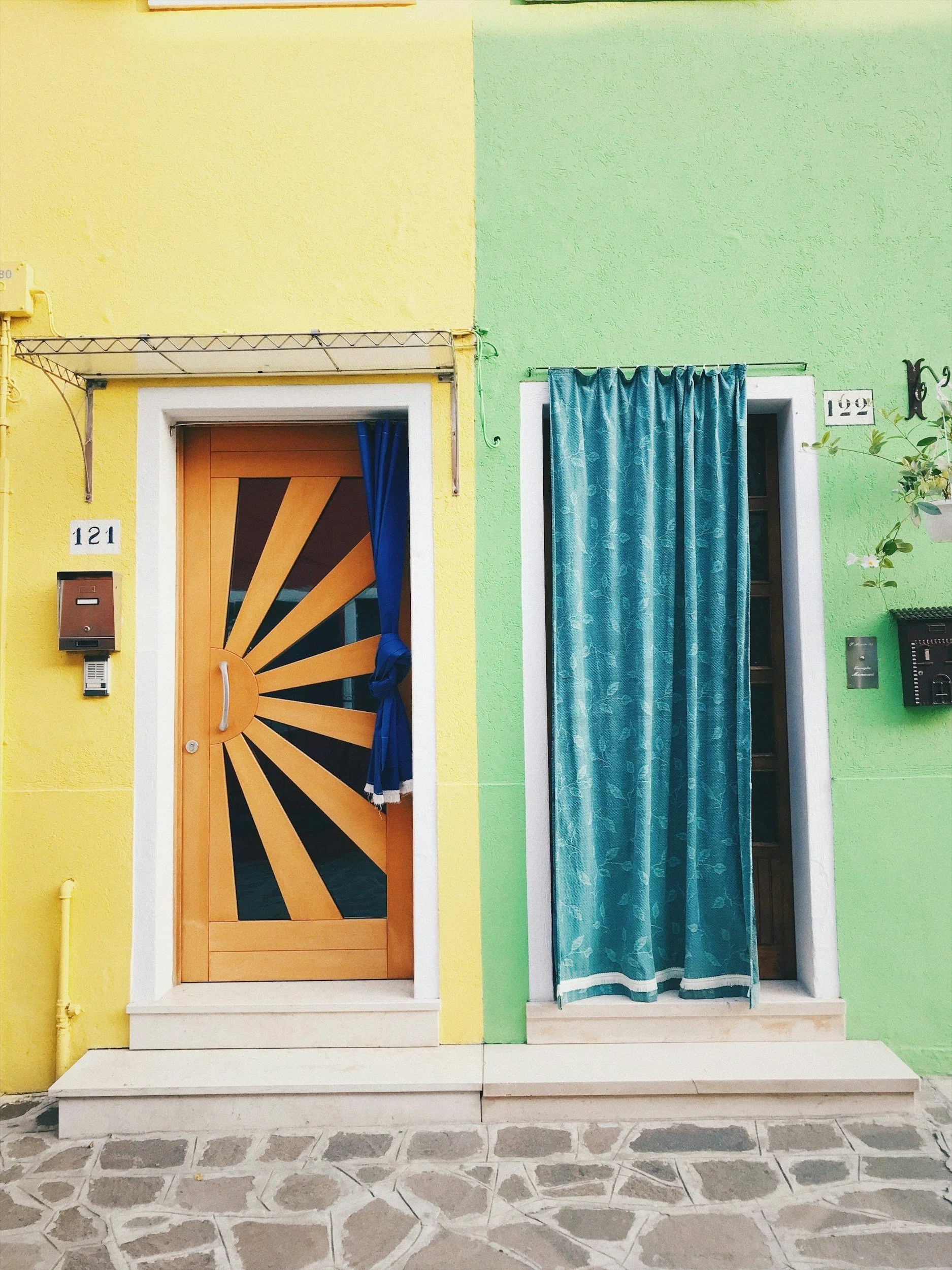 Front view of a colorful house with a yellow wall on the left and a green wall on the right. The house has two doors, one with a sunburst design and a blue curtain on the left, and a window with a blue curtain on the right. House numbers '121' and '122' are visible above the doors. The ground has gray and white patterned tiles.