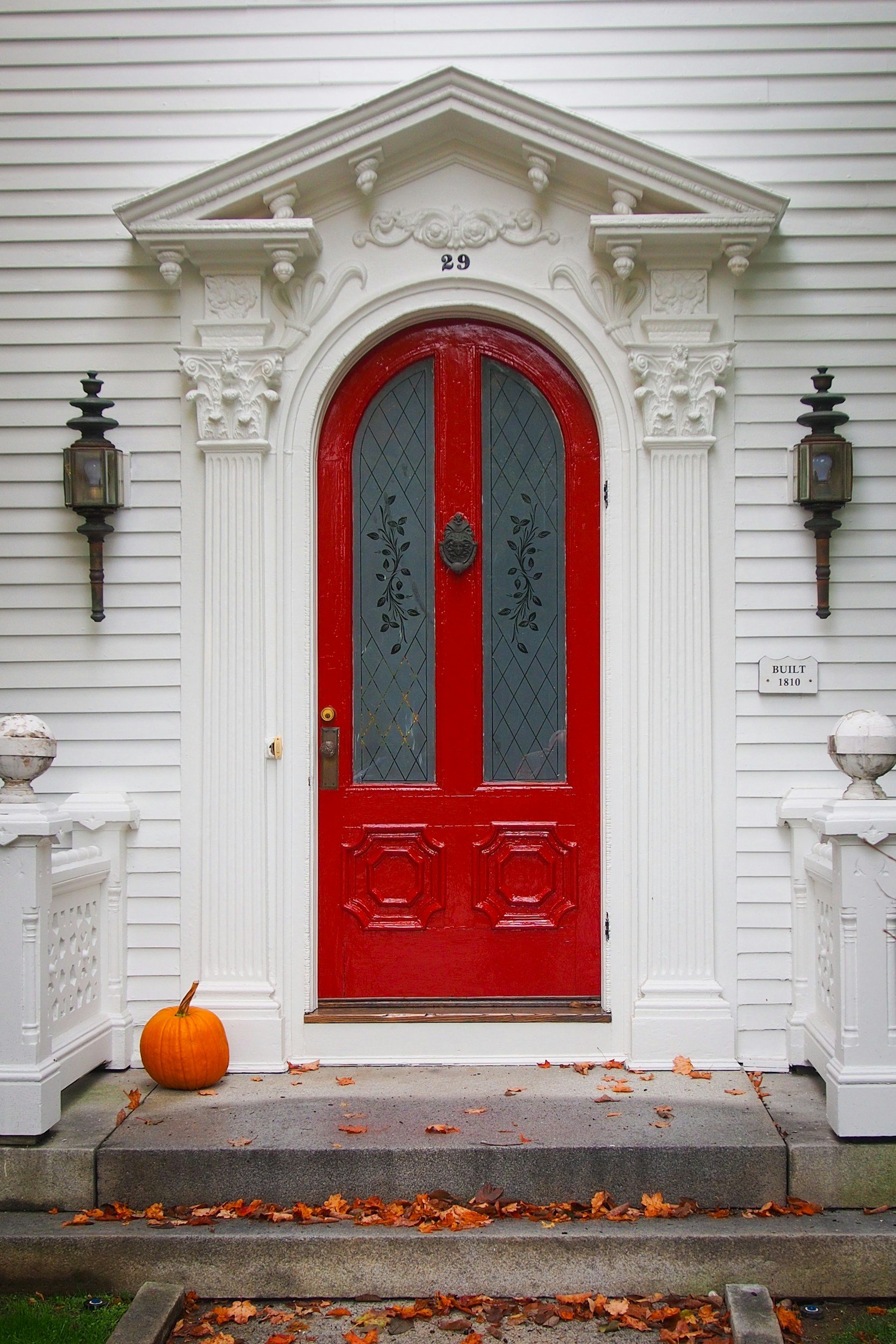 Front porch of a house with a red door, white decorative trim, black lantern lights on each side, a pumpkin on the left, and fallen autumn leaves on the steps and ground.