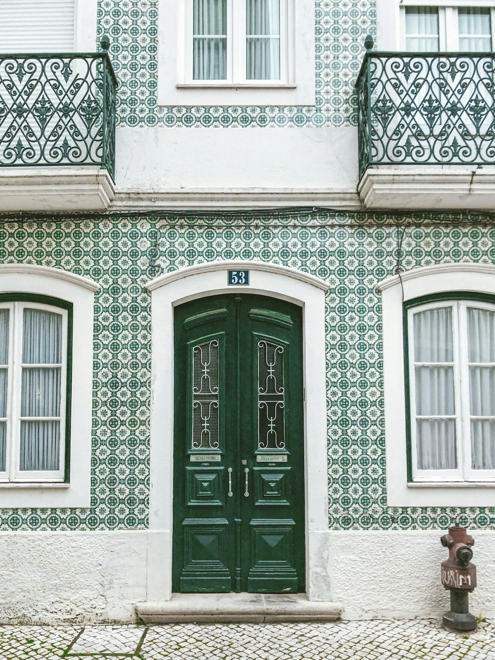 The front entrance of a building with green double doors and decorative metal grilles. The facade is covered with patterned tiles in green and white, and there are four windows with white frames and curtains. A house number "53" is above the door, and a fire hydrant is on the sidewalk to the right.