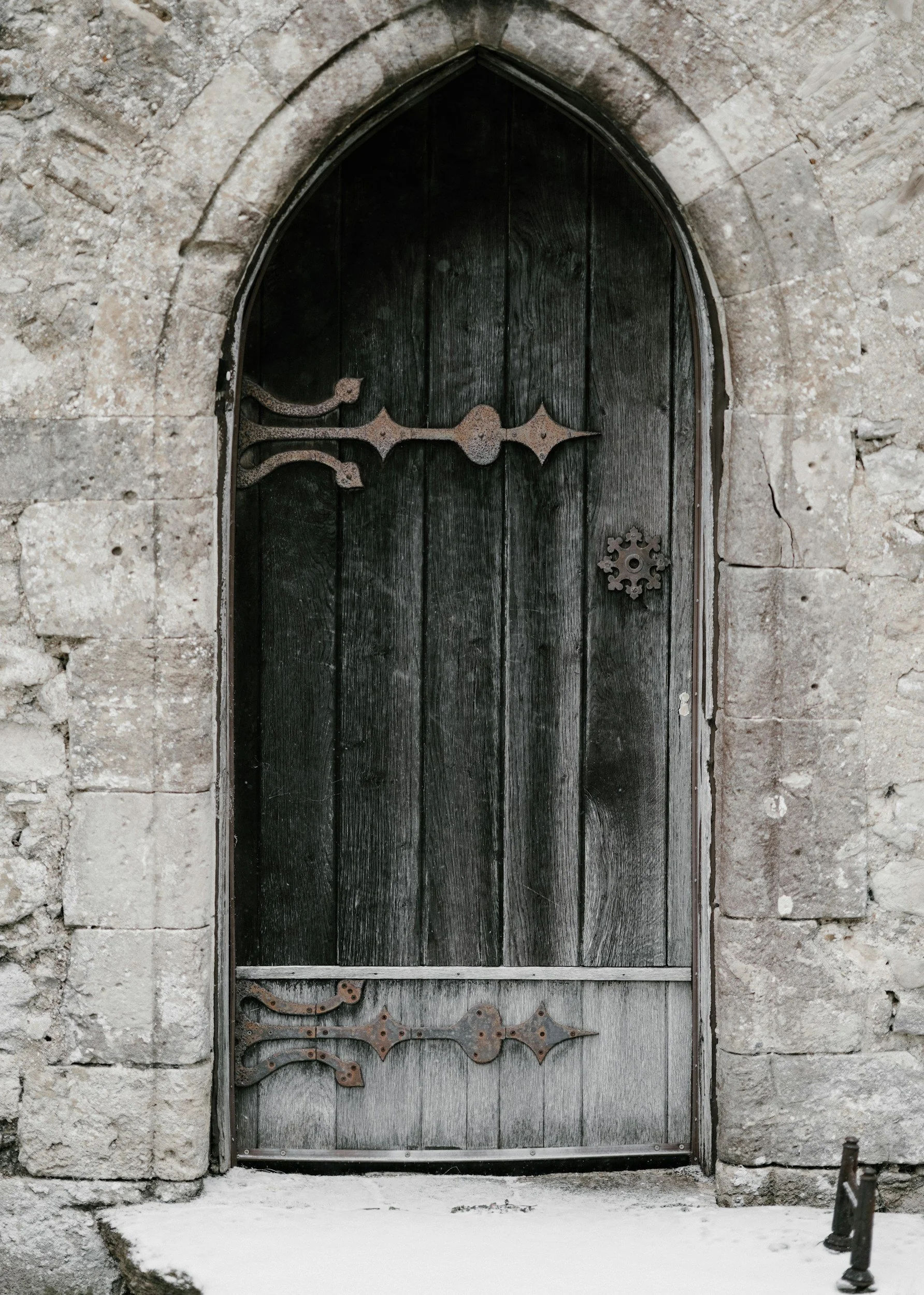 A dark wooden door with metal decorative hinges and gears, set within a stone archway with a rough stone wall surrounding it.