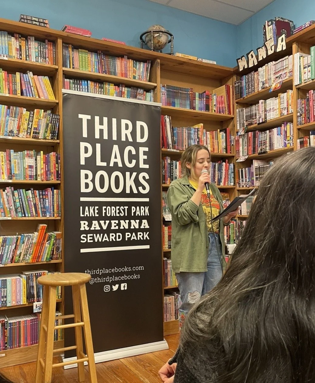 A woman speaking into a microphone while holding a paper in a bookstore with shelves of colorful books. A black banner reads 'Third Place Books' and lists locations: Lake Forest Park, Ravenna, Seward Park. Part of a small audience is visible in the foreground.
