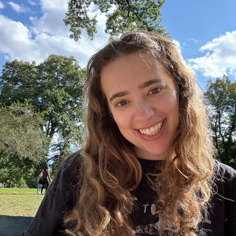 Smiling young woman with long, wavy hair outdoors in a park, with trees and blue sky in the background.