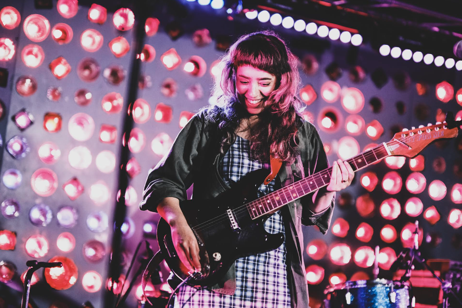 A woman with wavy hair and bangs, smiling while playing an electric guitar on stage with colorful circular lights in the background.