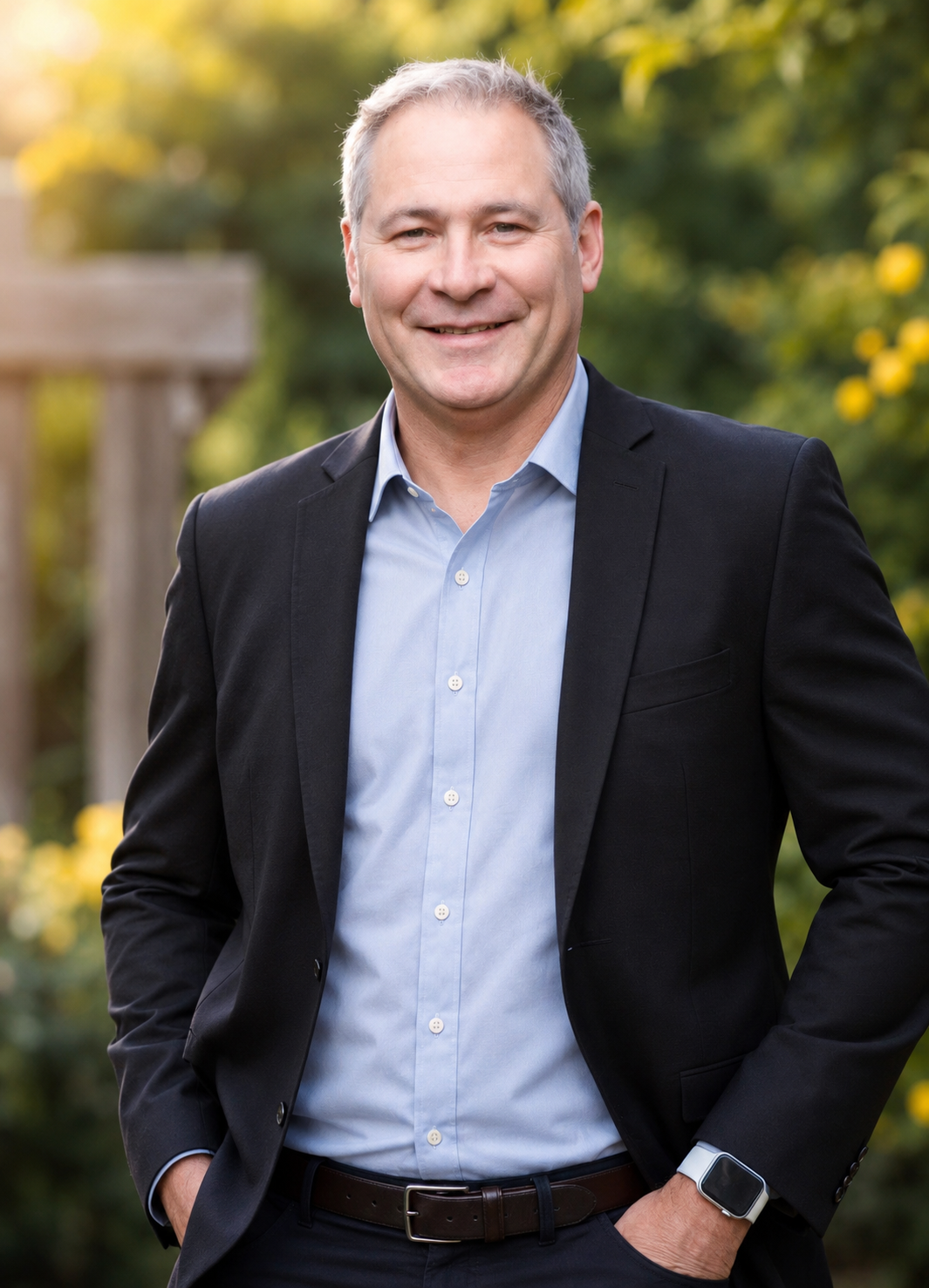 A middle-aged man in a black suit jacket and light blue dress shirt standing outdoors with a blurred background of green foliage and yellow flowers, smiling at the camera.
