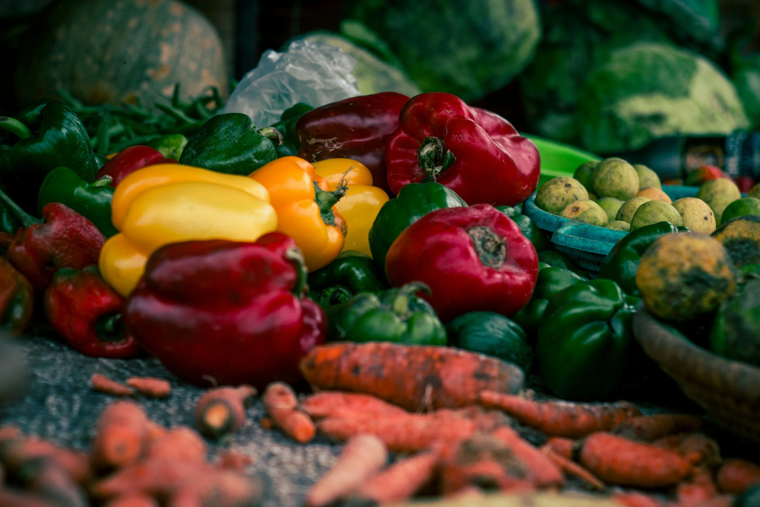 A variety of fresh colorful peppers and vegetables on a market stall, including green, red, yellow, and orange peppers, green and yellow tomatoes, and carrots.