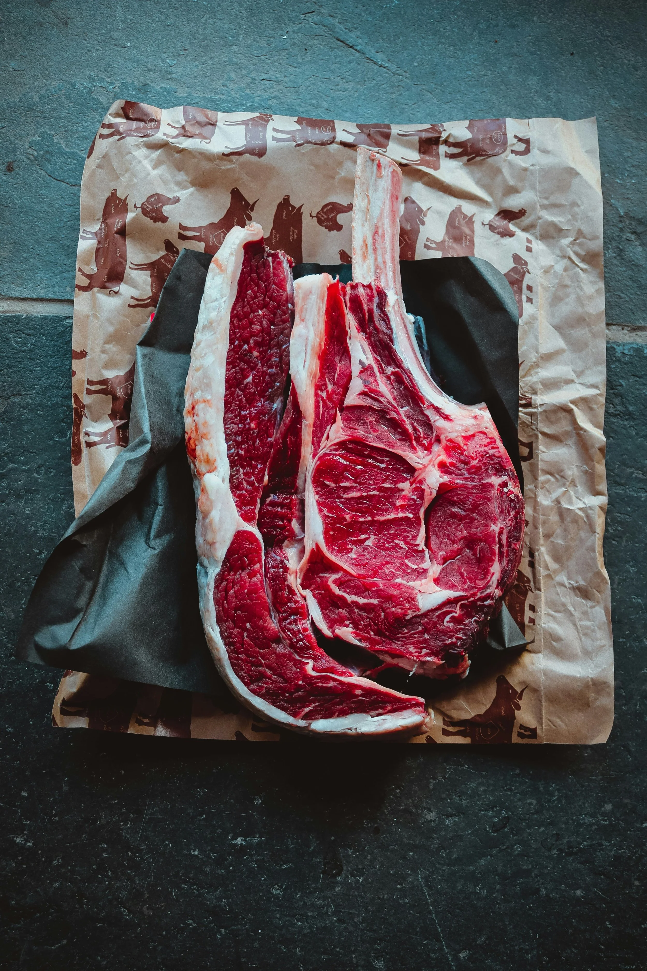 A large, raw beef ribeye steak on butcher paper on a dark countertop.