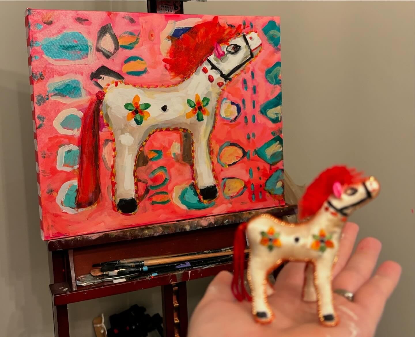 A hand holding a small decorated horse figurine in front of an abstract painting of a similar horse on an easel. The painting features a white horse with black details, red mane, and floral decorations on its body, set against a pink background with 
