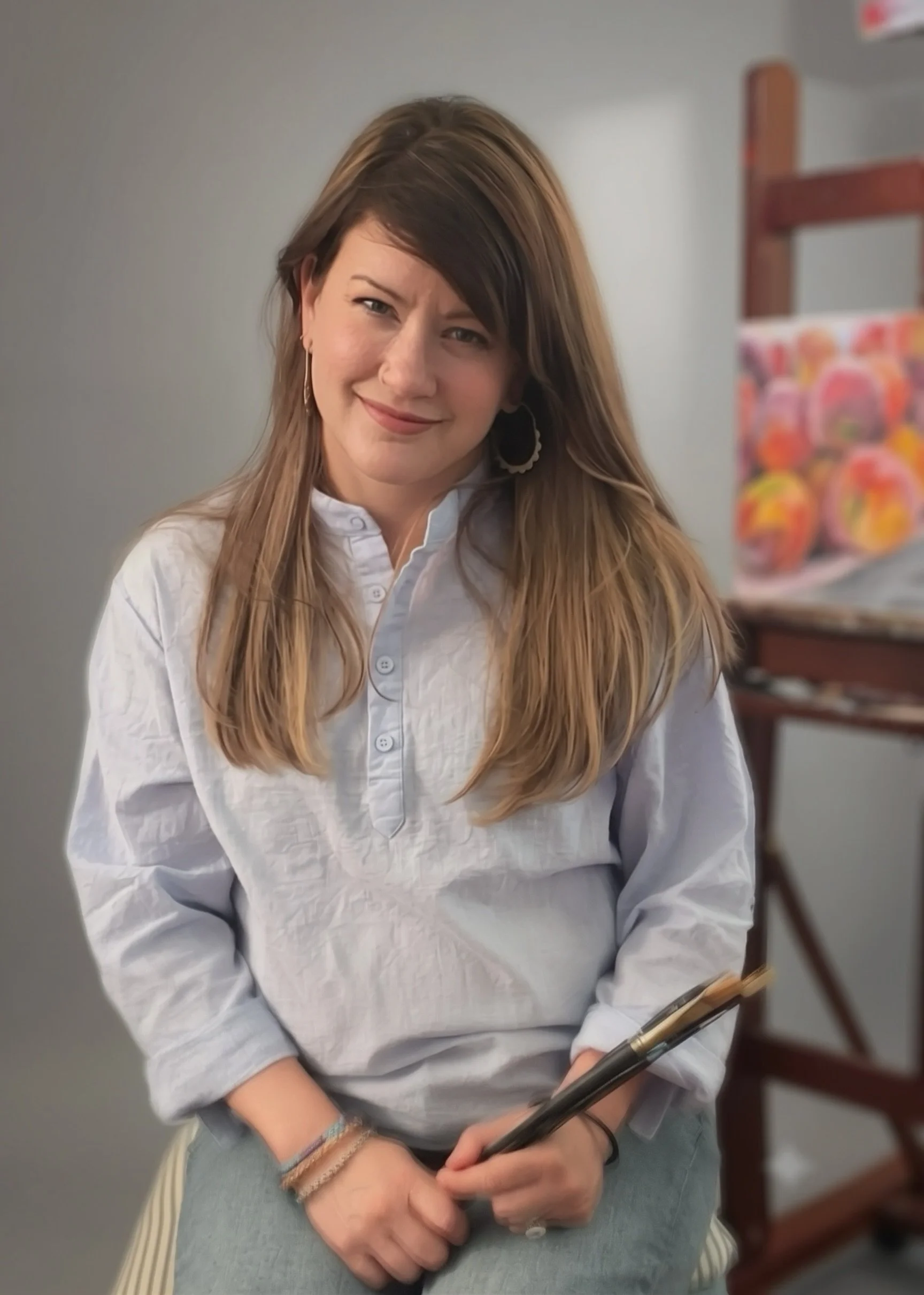 A woman with long brown hair, wearing a blue linen button-up shirt and earrings, sitting at a table with her hands clasped holding a paintbrush with a painting in the background.
