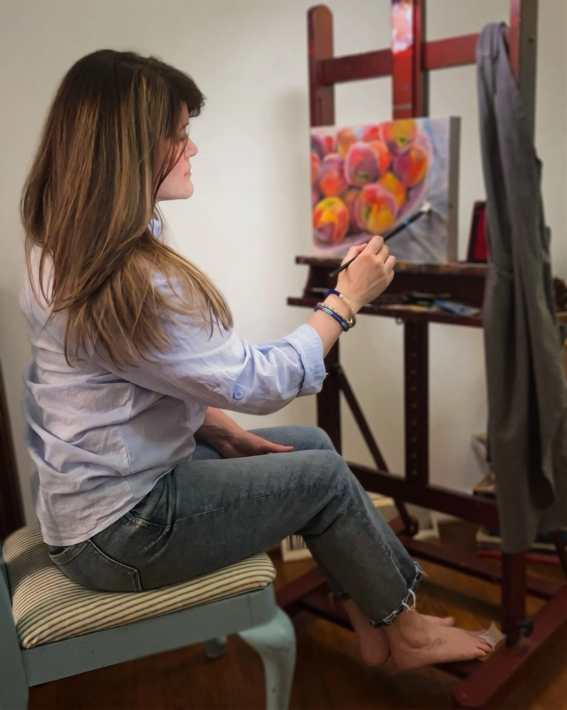 A woman painting on a canvas in an art studio, sitting on a striped cushioned chair with her legs crossed, wearing a white shirt and jeans.