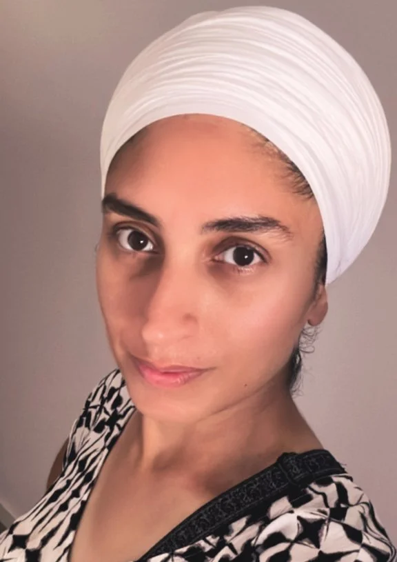A woman wearing a white turban and a black and white patterned top, looking at the camera with a neutral expression.