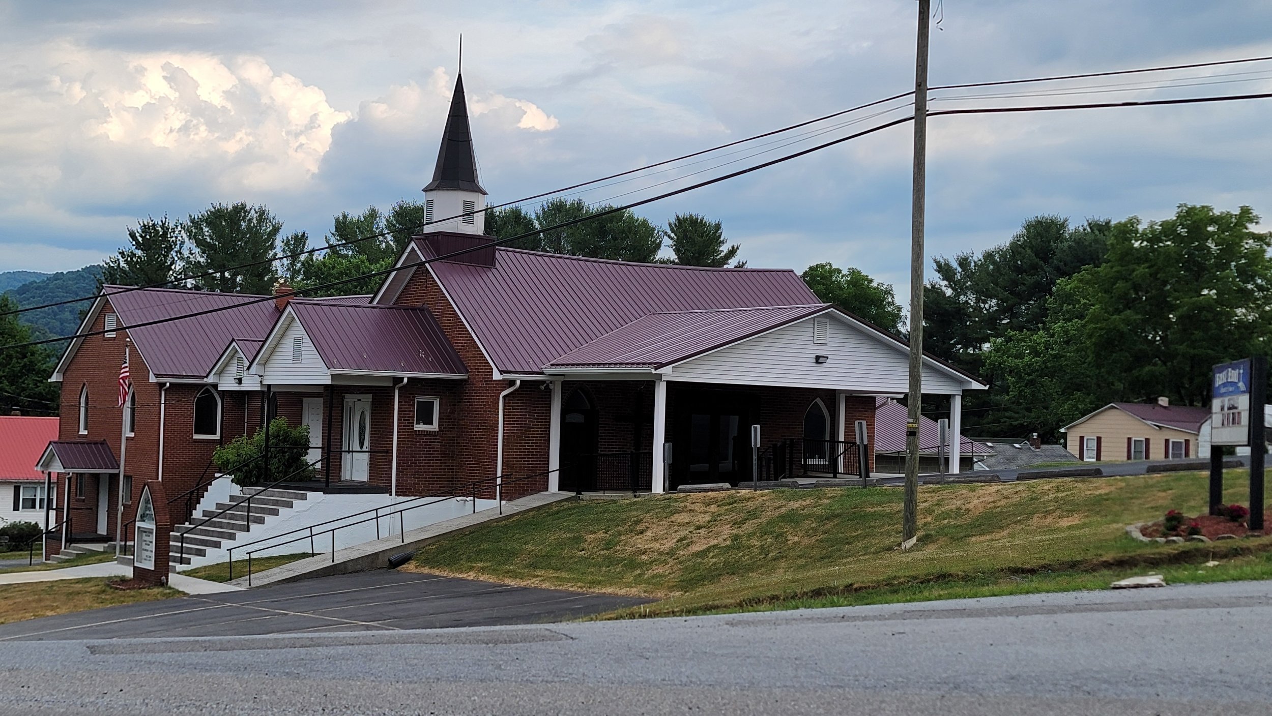 A brick church with a steeple, white trim, and a red metal roof, located on a grassy hill with a parking lot in the foreground, surrounded by trees and other small buildings, with a cloudy sky overhead.