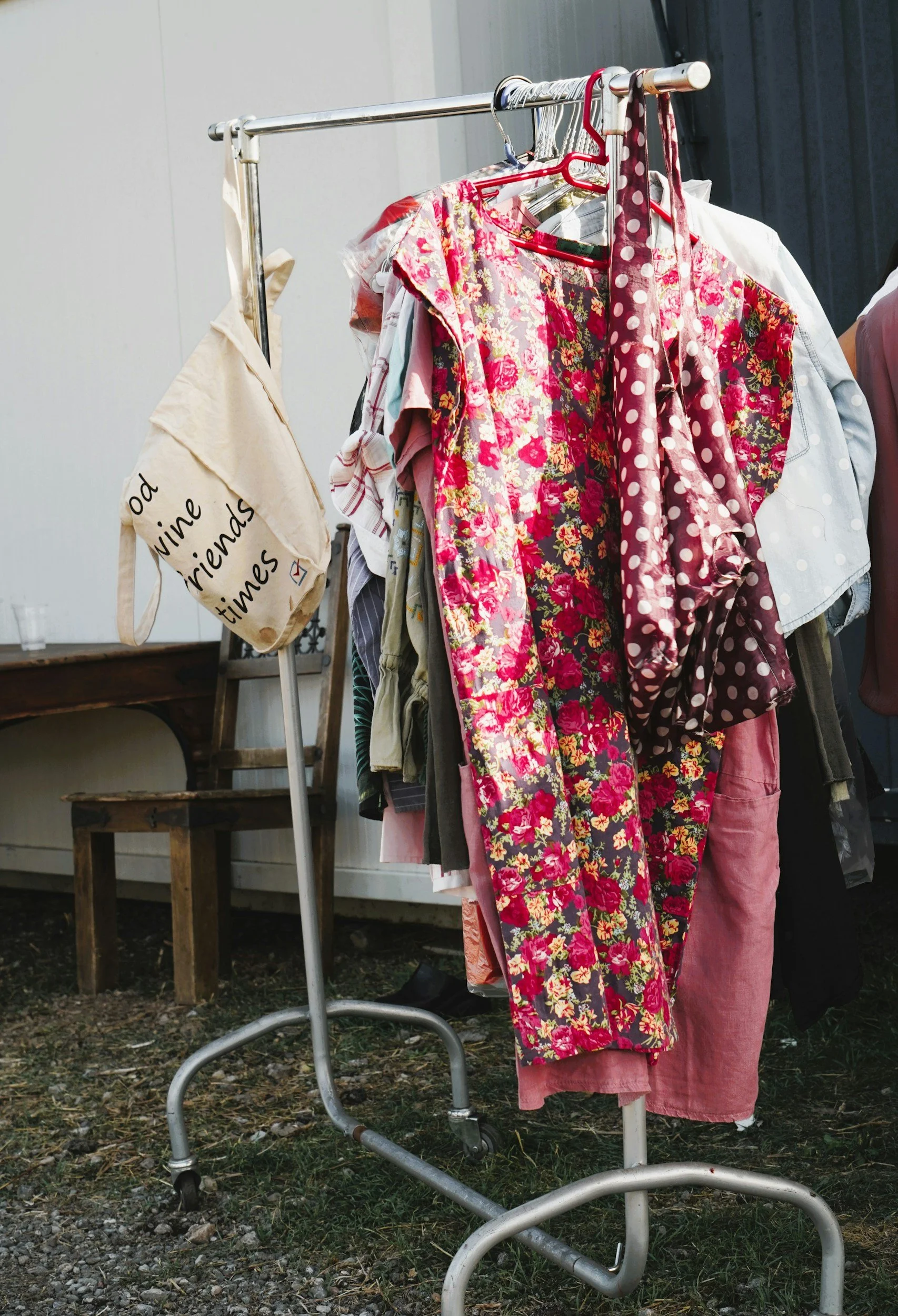 Clothing on a metal rack with wheels, including floral dresses, pink pants, and various other garments, outside on a patch of grass and gravel.