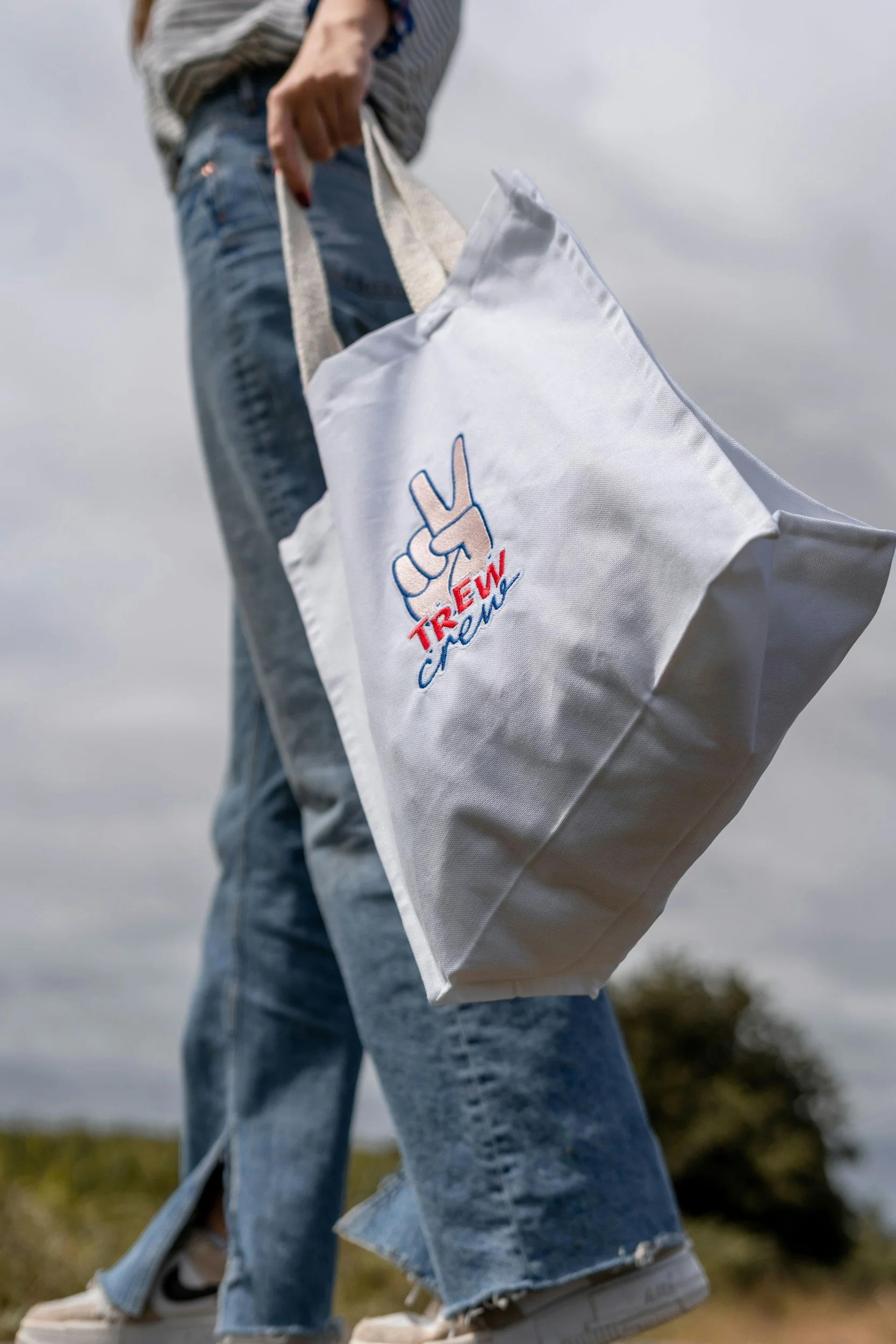 Person wearing jeans, sneakers, and a striped shirt, holding a white tote bag with the phrase 'Trieu crew' and a peace sign illustration, outdoors with cloudy sky background.