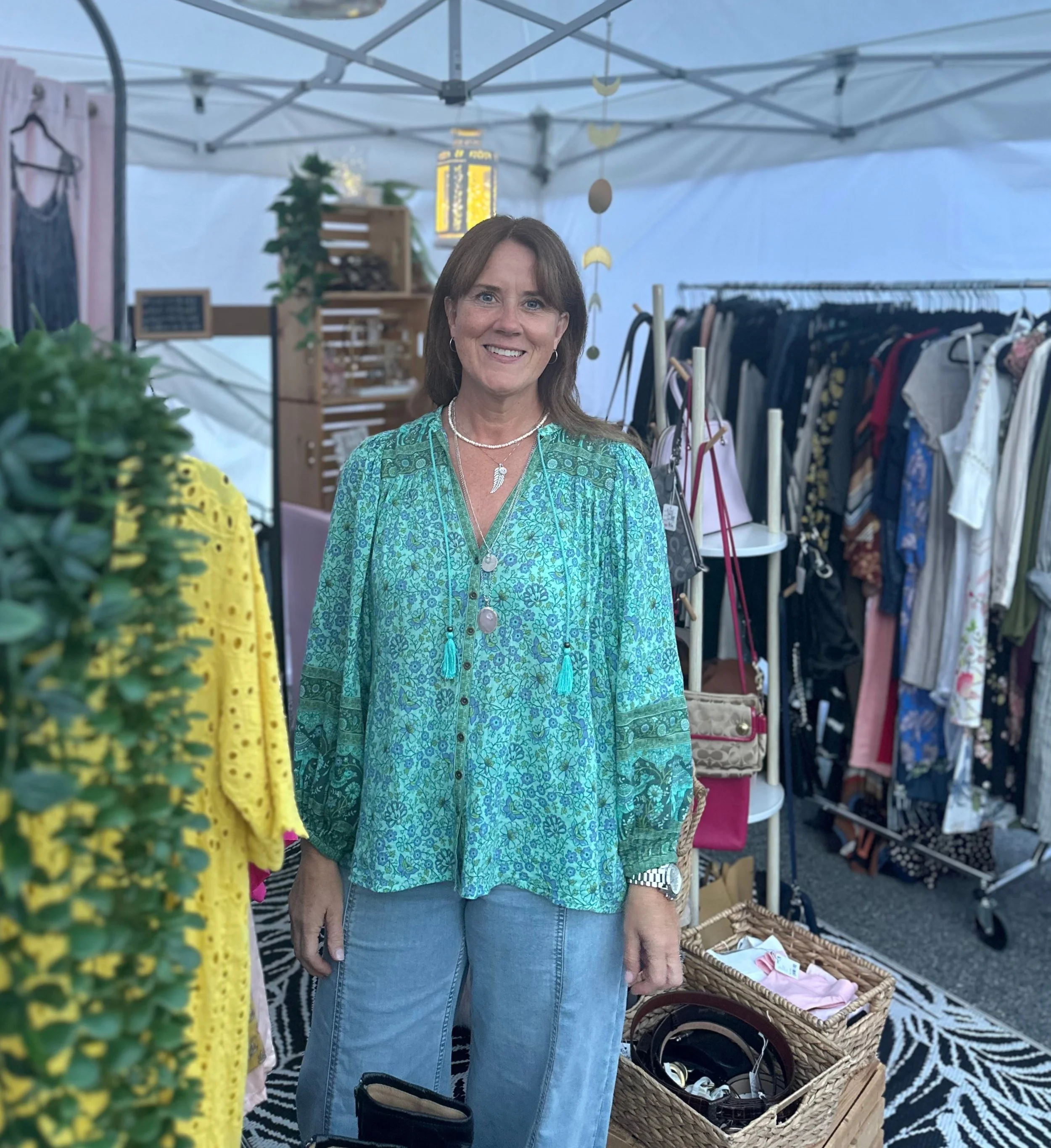 A woman with brown hair smiling, wearing a blue-green patterned blouse, a pearl necklace, and a feather necklace. She is standing inside a clothing and accessory booth at an outdoor market with racks of clothes and a wicker basket filled with folded clothes around her. The background shows a tent structure and decorative hanging moons and circles.