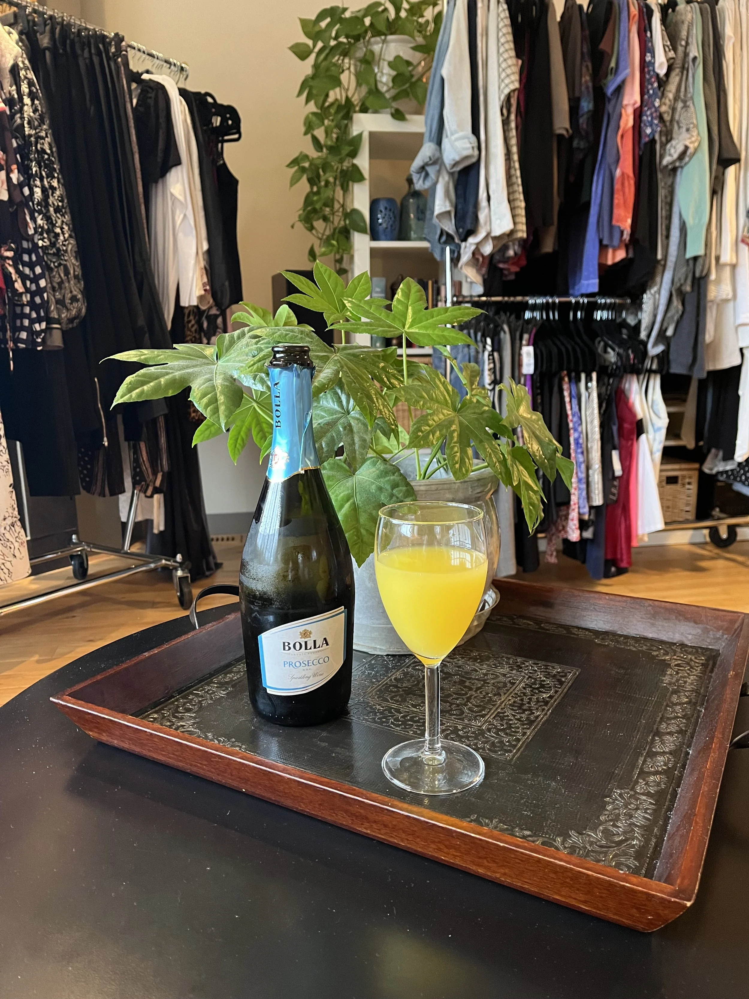 A glass of yellow drink and a bottle of Bolla prosecco on a wooden tray with a plant in the background, set on a black table in a closet or dressing room with clothing racks and shelves.