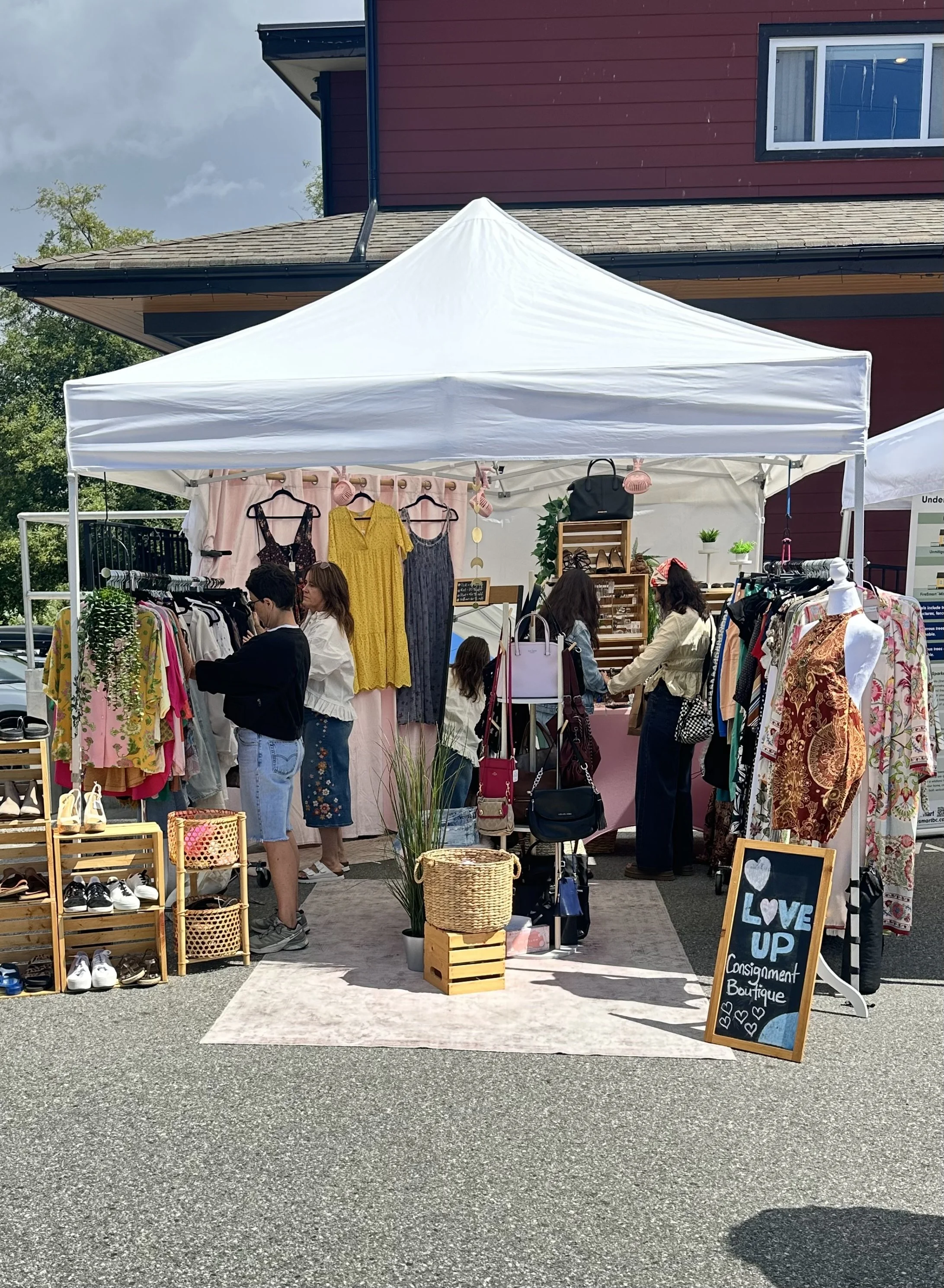 Outdoor boutique stall with clothing, shoes, and accessories on display. Several people browse the items under a white canopy, with a red building and blue sky in the background.