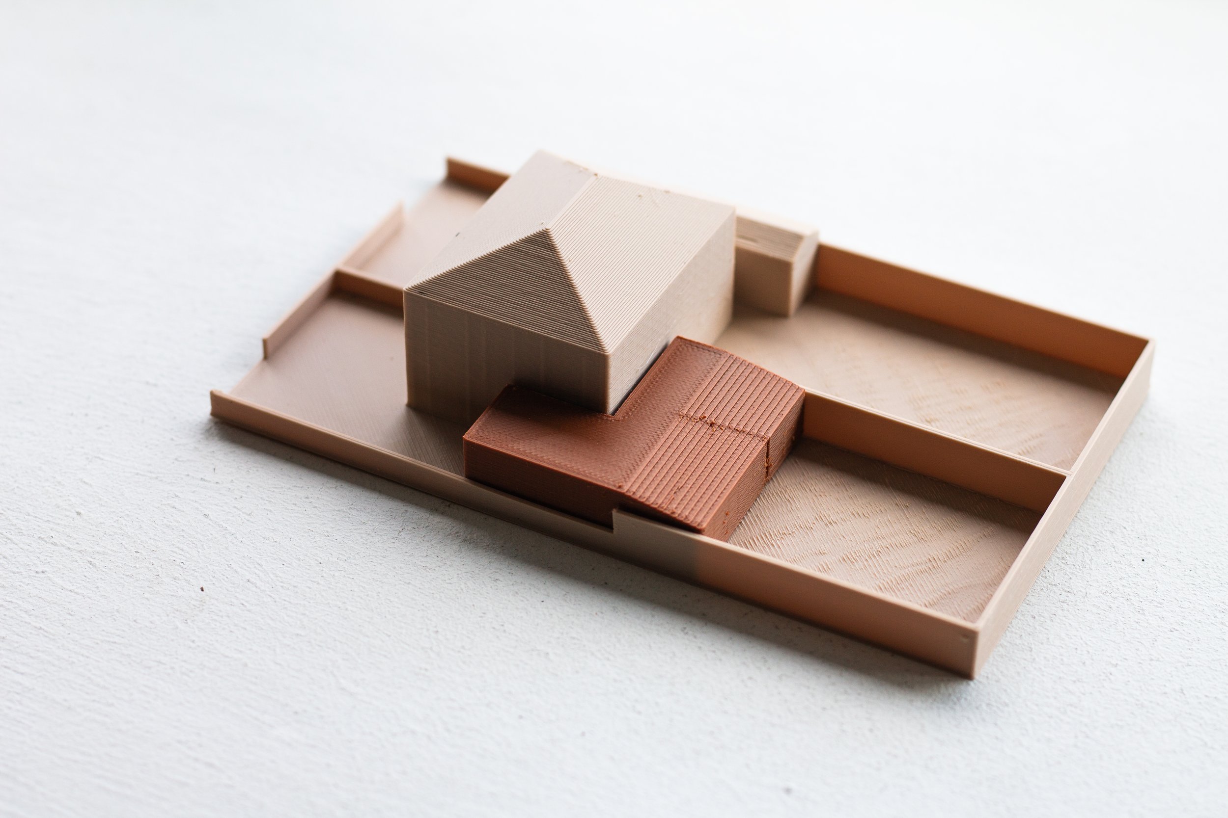 Architectural model of a house with a triangular roof, made of textured material, placed on a rectangular base with surrounding framing.