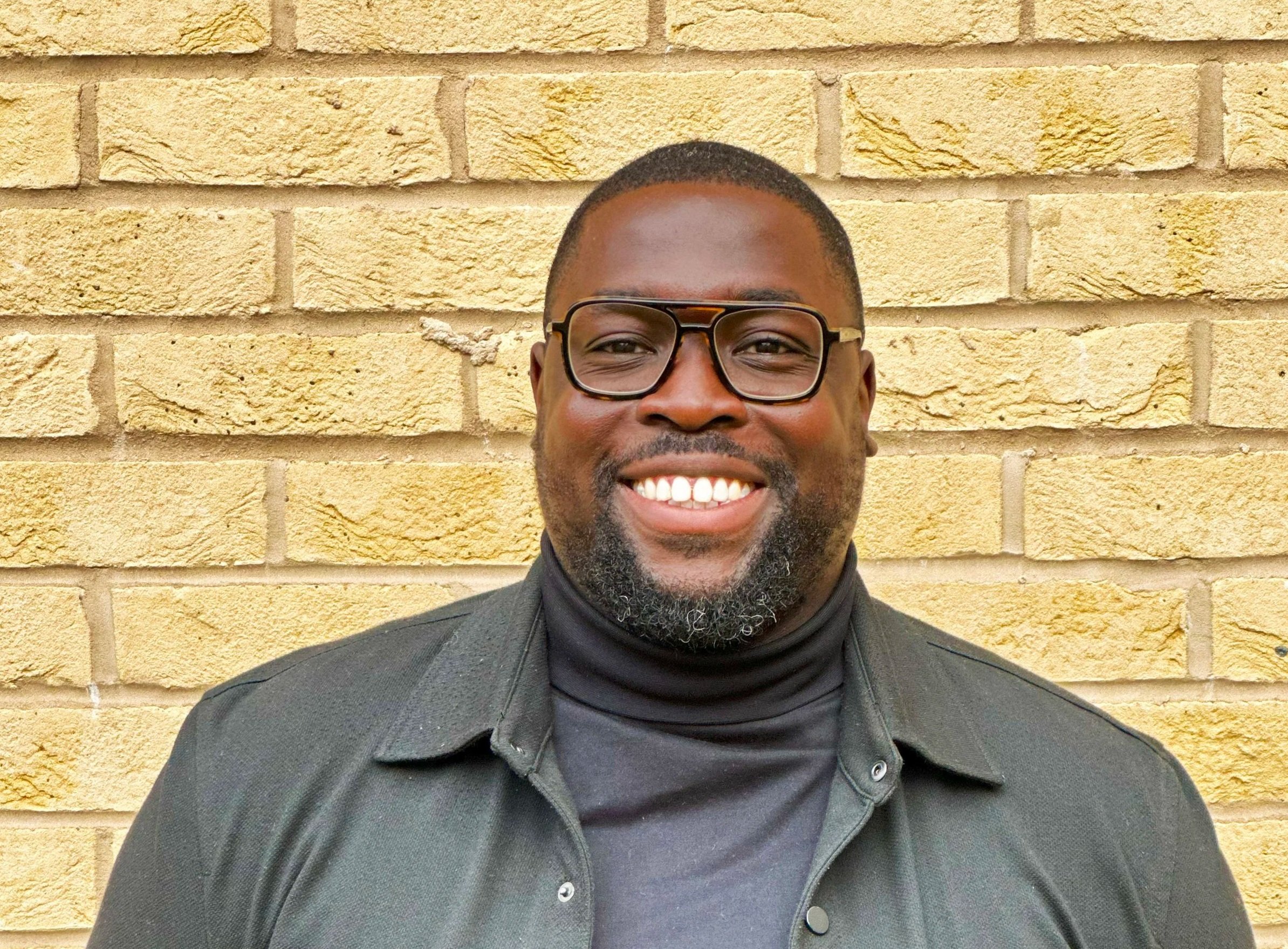 Close-up of smiling man wearing glasses and black jacket standing against yellow brick wall.