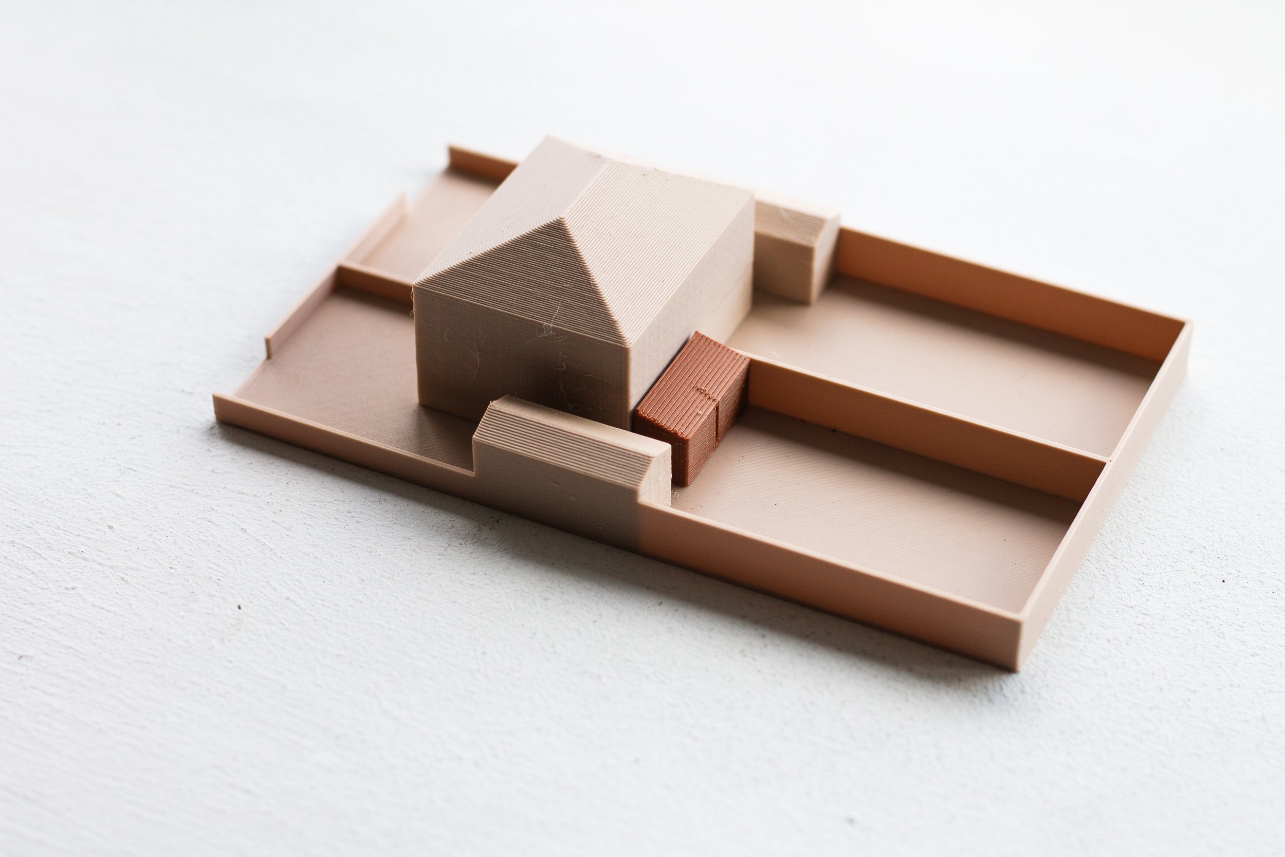 A minimalistic architectural model of a house with a sloped roof, set on a rectangular base, made of beige, brown, and terracotta colored materials.