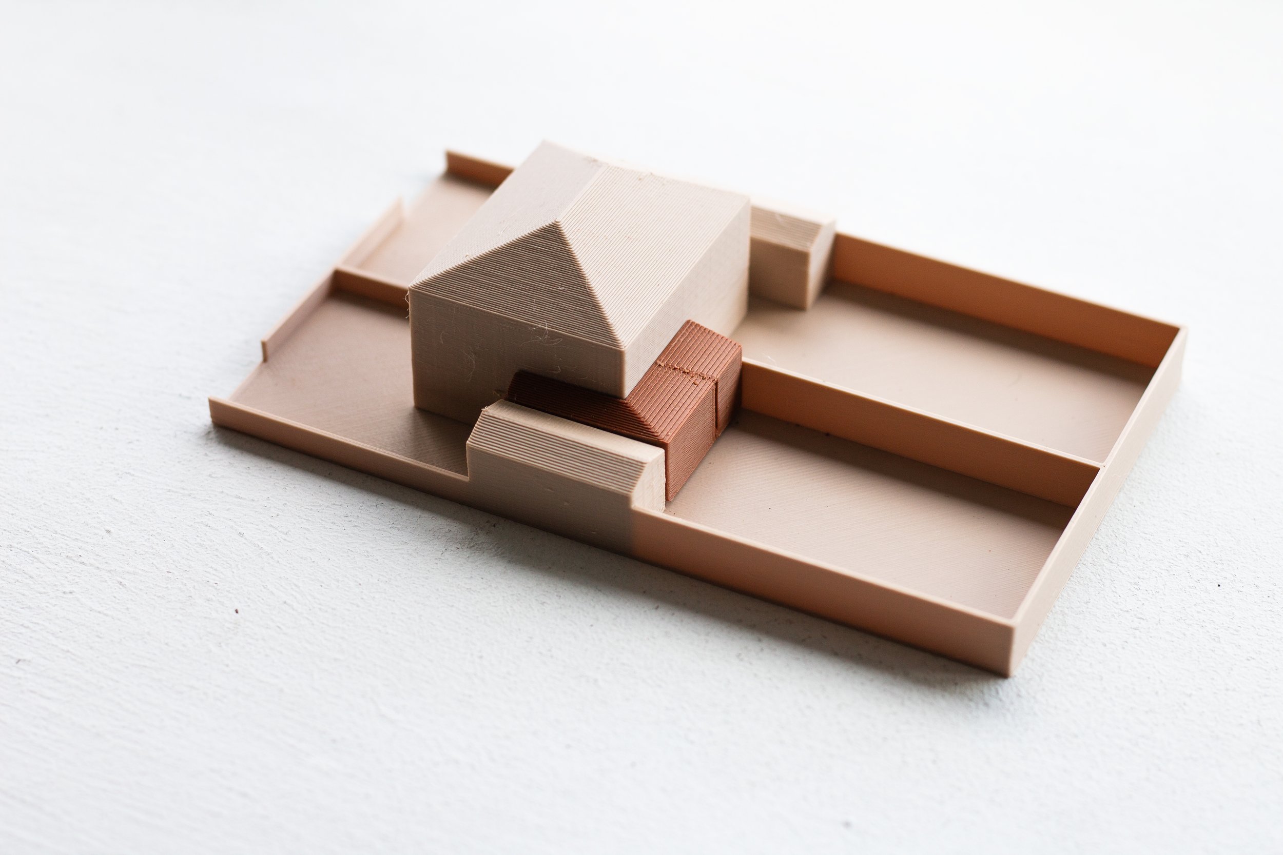 A minimalist architectural model featuring a small house with a gabled roof, set on a grid-like base with surrounding walls, all in neutral tones.