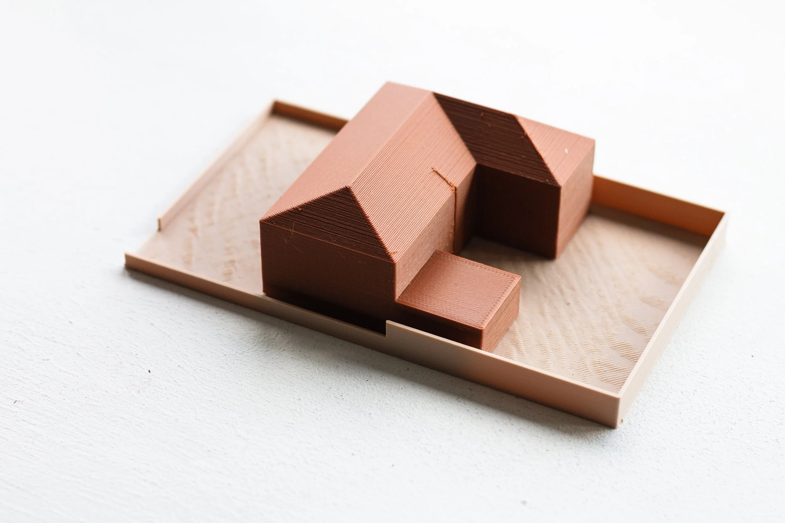 A small architectural model of a house made from pinkish-brown material, placed on a light-colored wooden tray.