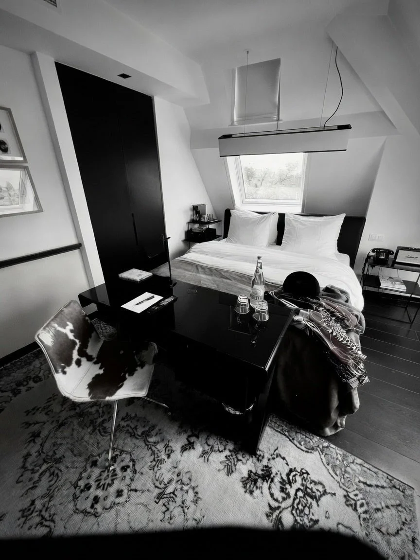 Bedroom with a bed under a sloped ceiling, a window behind the headboard, a black wardrobe, a table with a bottle of water and glasses, a chair with cow-print upholstery, and a patterned rug.