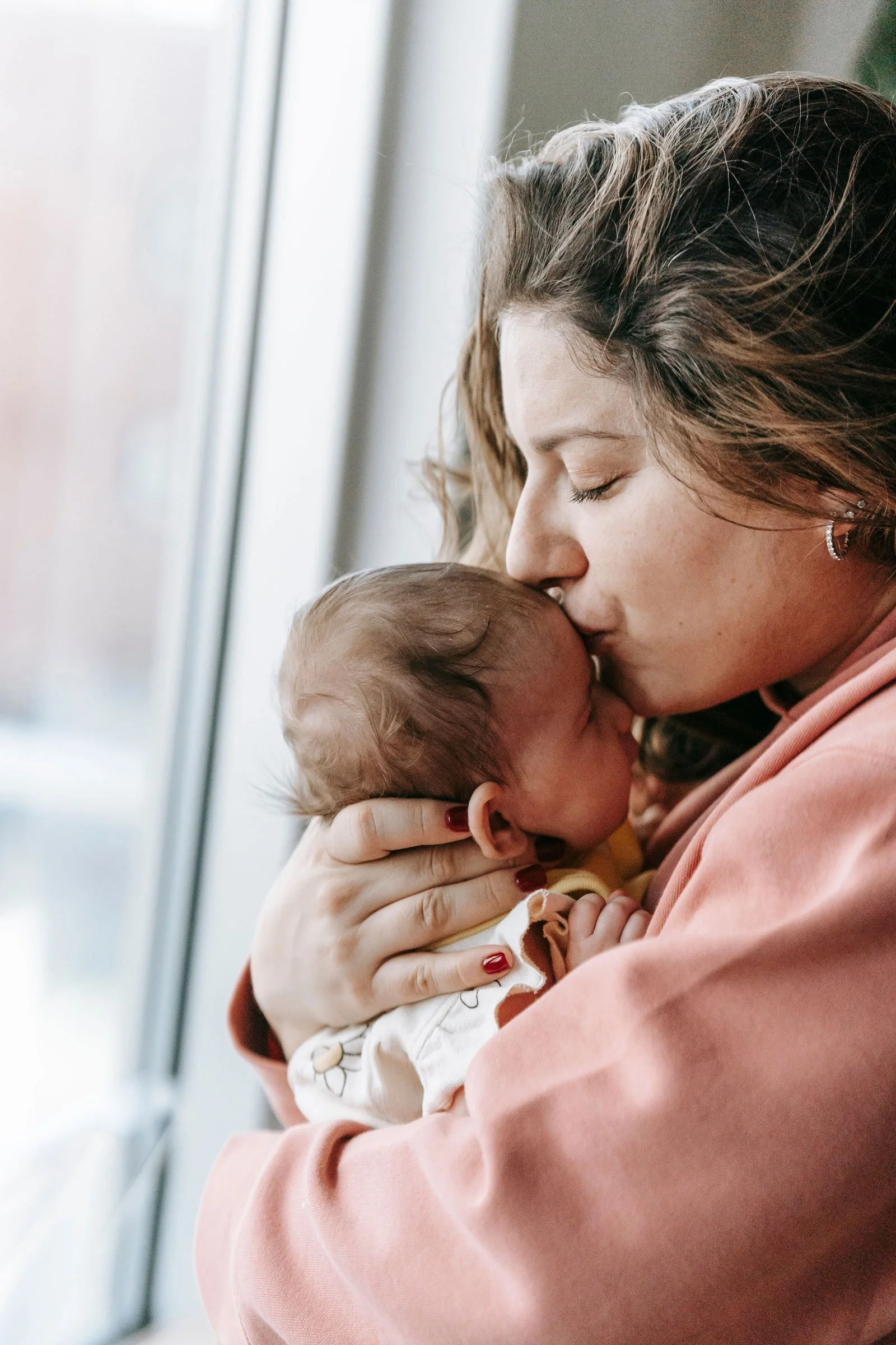 Une femme avec des cheveux bruns foncés et un maquillage léger donne un câlin tendre à un bébé, en fermant les yeux, près d'une fenêtre, dans un intérieur doux et chaleureux.