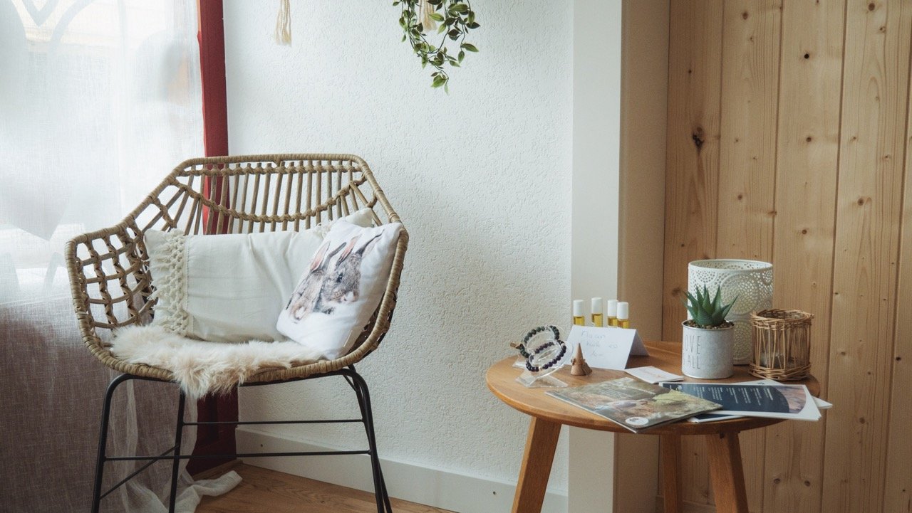 Coin salon avec un fauteuil en osier, un coussin à motif de renard, un plaid en fourrure blanche, et une petite table en bois avec des magazines, une plante en pot, des bouteilles de produits cosmétiques et autres objets décoratifs.