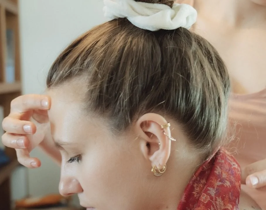 Une jeune femme avec des cheveux attachés en chignon, portant plusieurs piercings à l'oreille, reçoit un soin de quelqu'un.