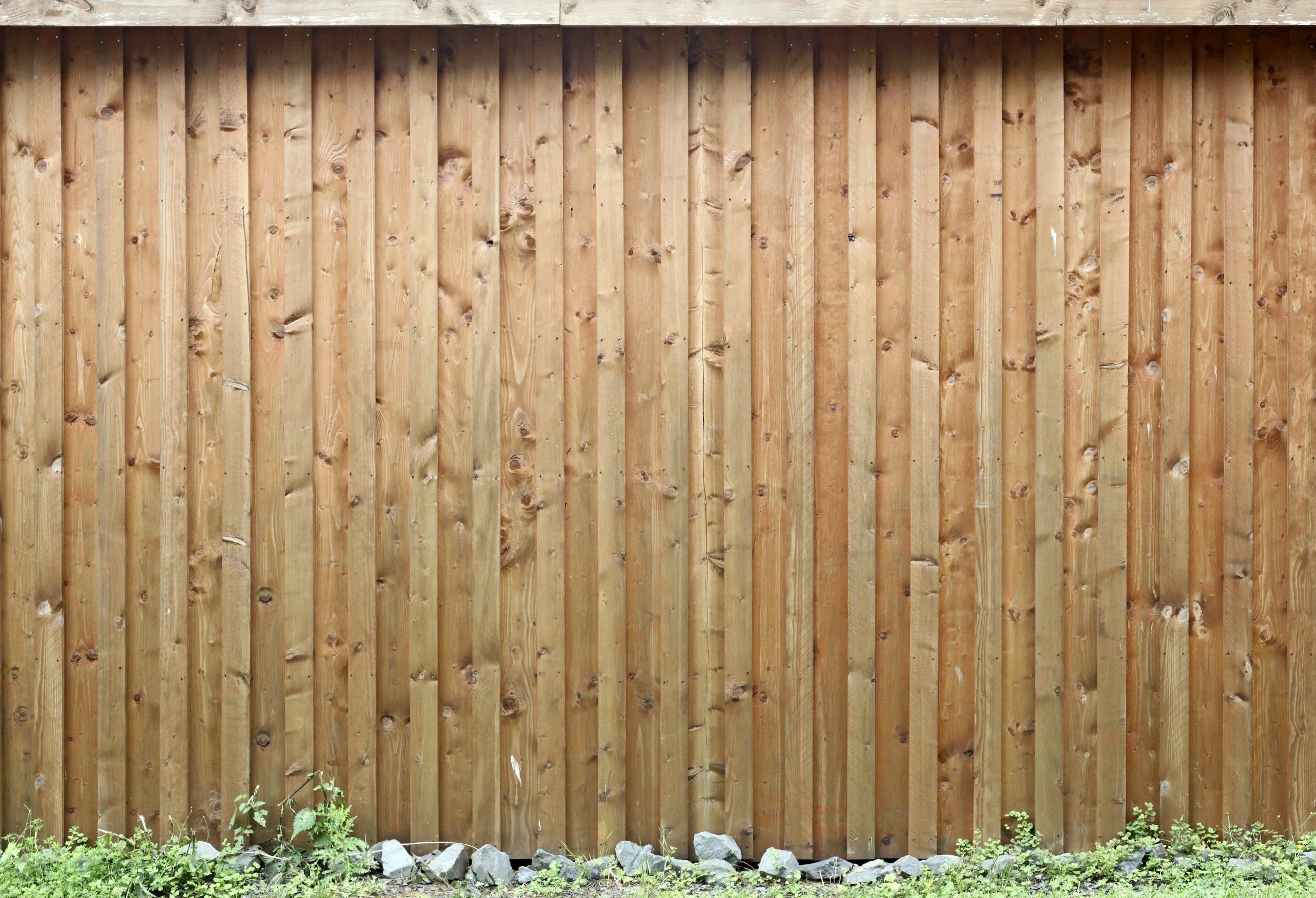A wooden privacy fence with vertical planks and a horizontal top rail, with some green plants and rocks at the base.