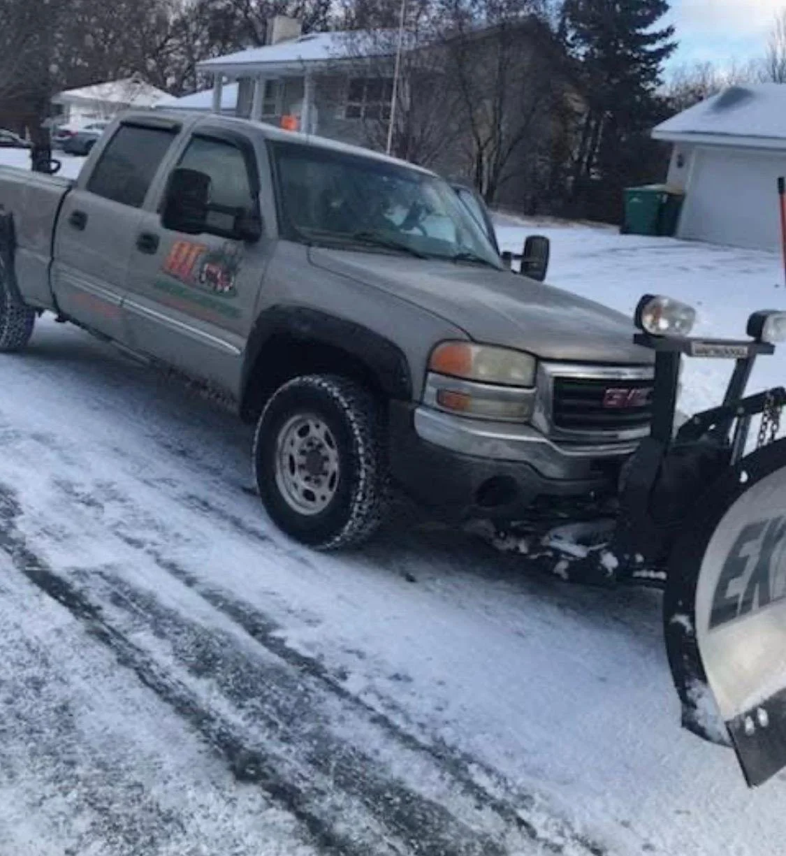 A gray GMC pickup truck attached to a snowplow clearing a snow-covered driveway on a winter day.