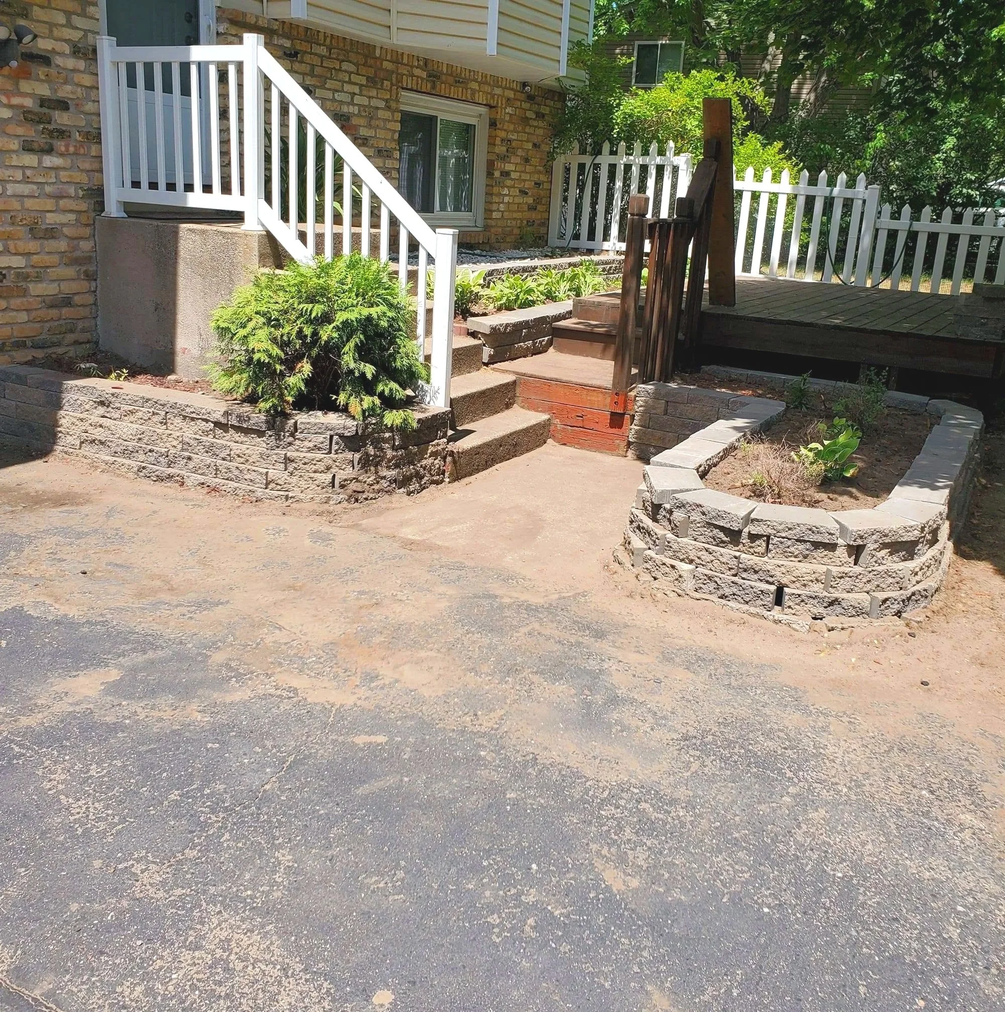 A backyard with a porch, a white picket fence, and some green plants. There are stairs leading up to the porch and trees in the background.