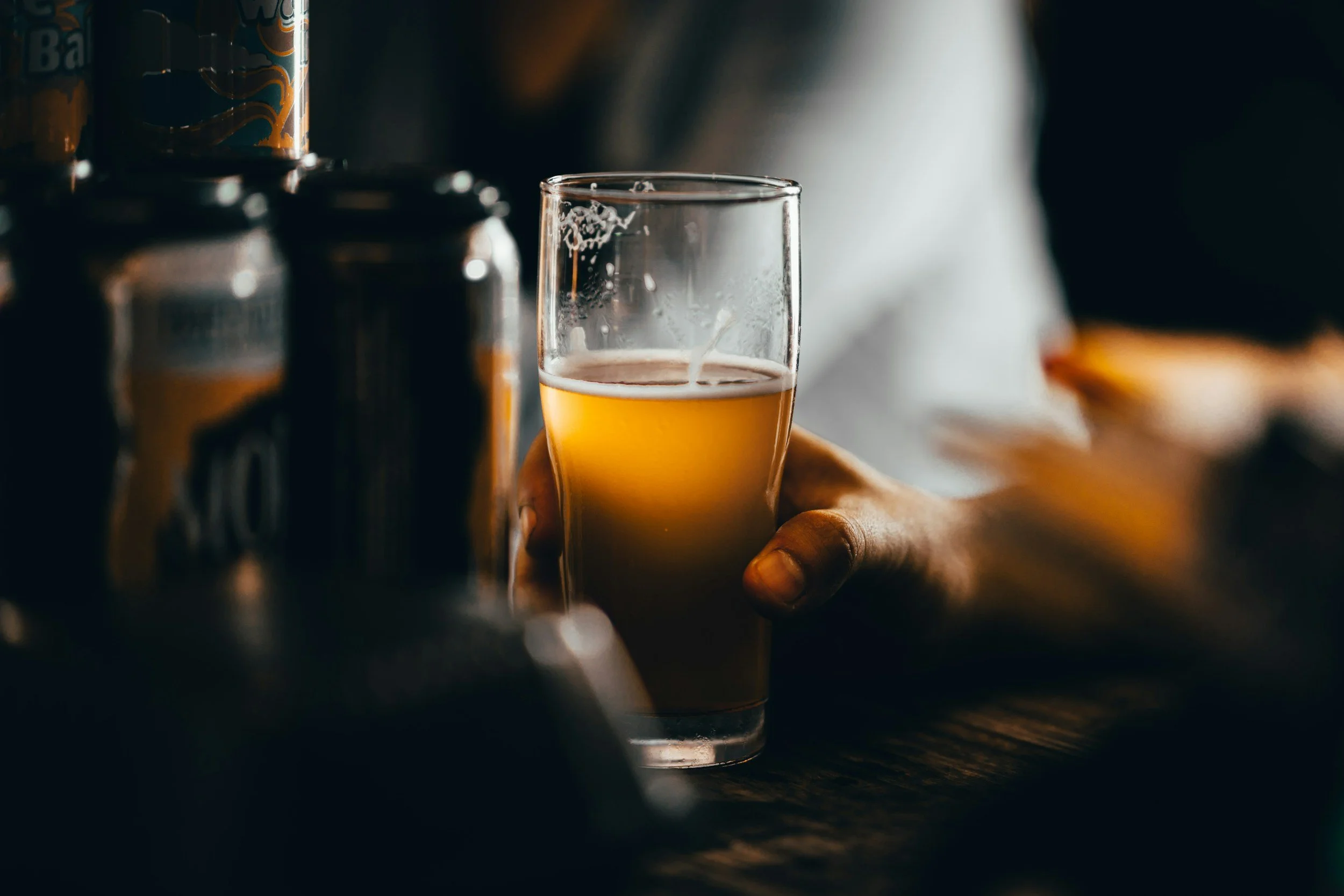 A close-up of a person’s hand holding a glass of beer, with cans of beer in the background. The scene appears to be in a dimly lit setting.